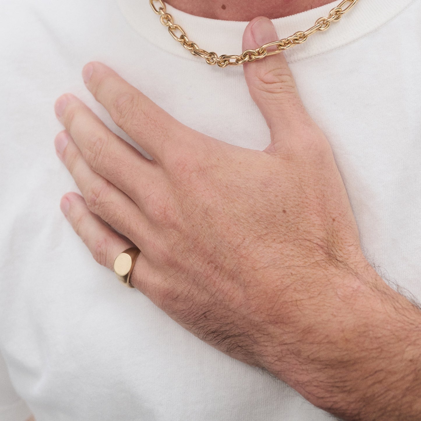 Man wearing a gold chain necklace and matching ring, resting hand on chest. White T-shirt enhances the luxury jewellery's elegance.