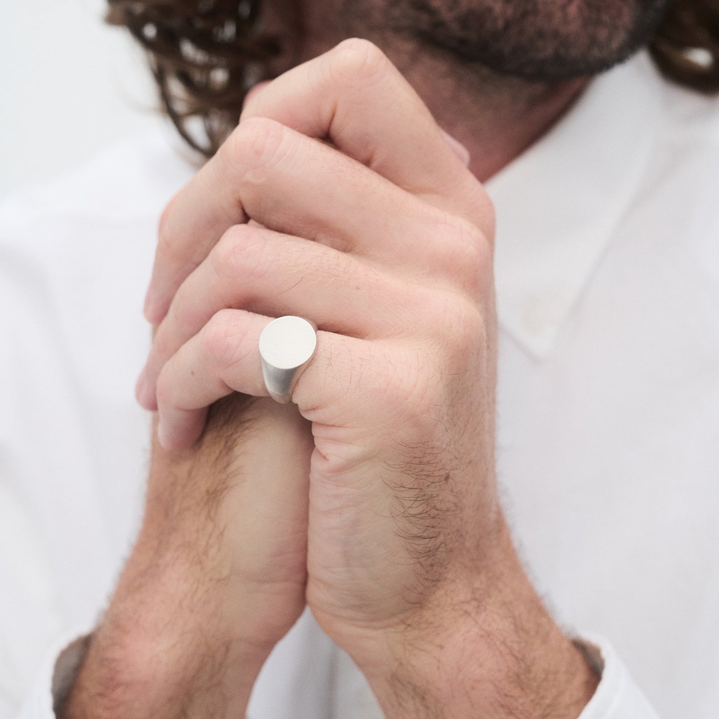 Close-up of a person wearing a minimalist silver ring, hands clasped. The person wears a white shirt, suggesting a stylish, refined look.