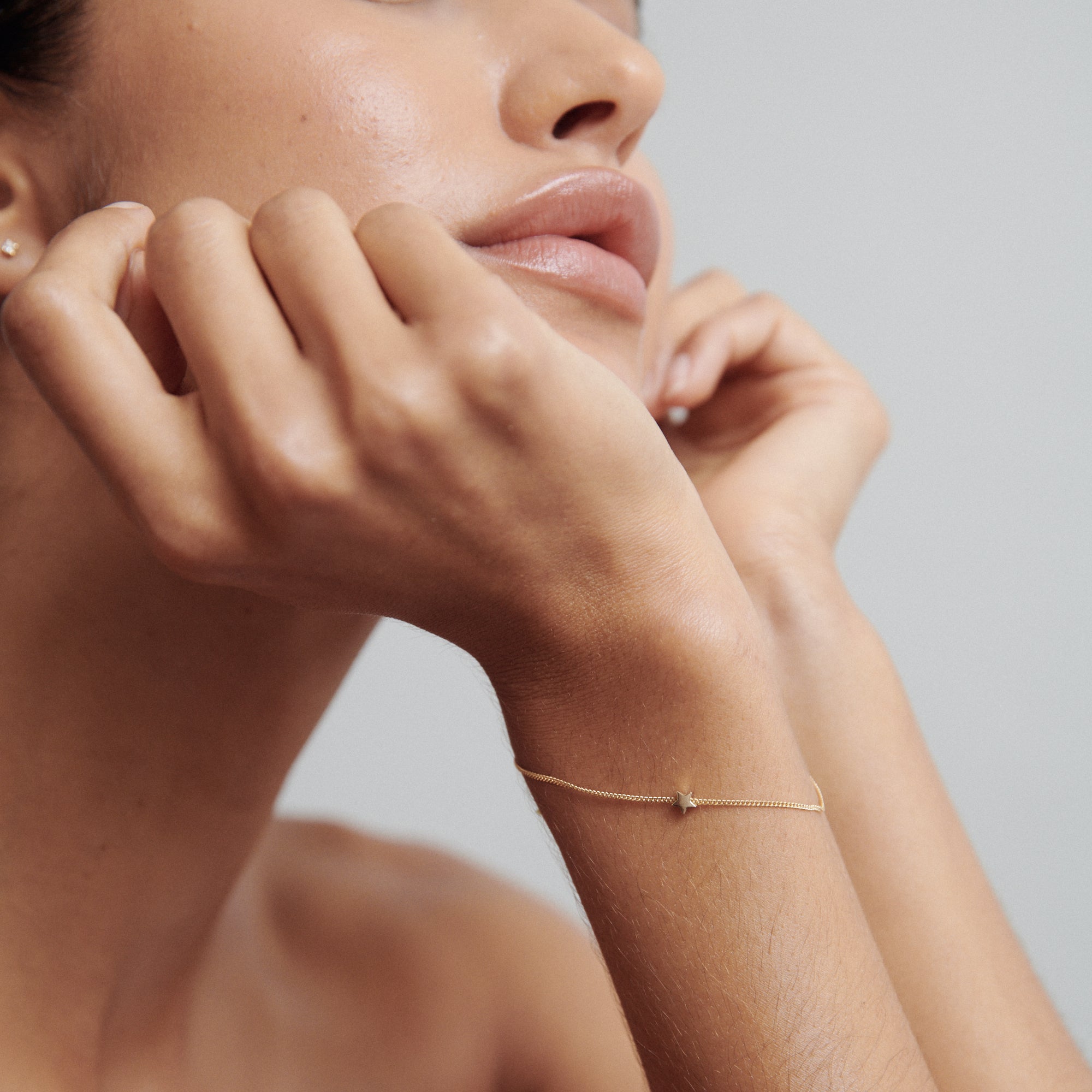 Woman posing with a minimalist gold bracelet, showcasing luxury jewellery. Close-up on her chin and hands against a soft grey background.