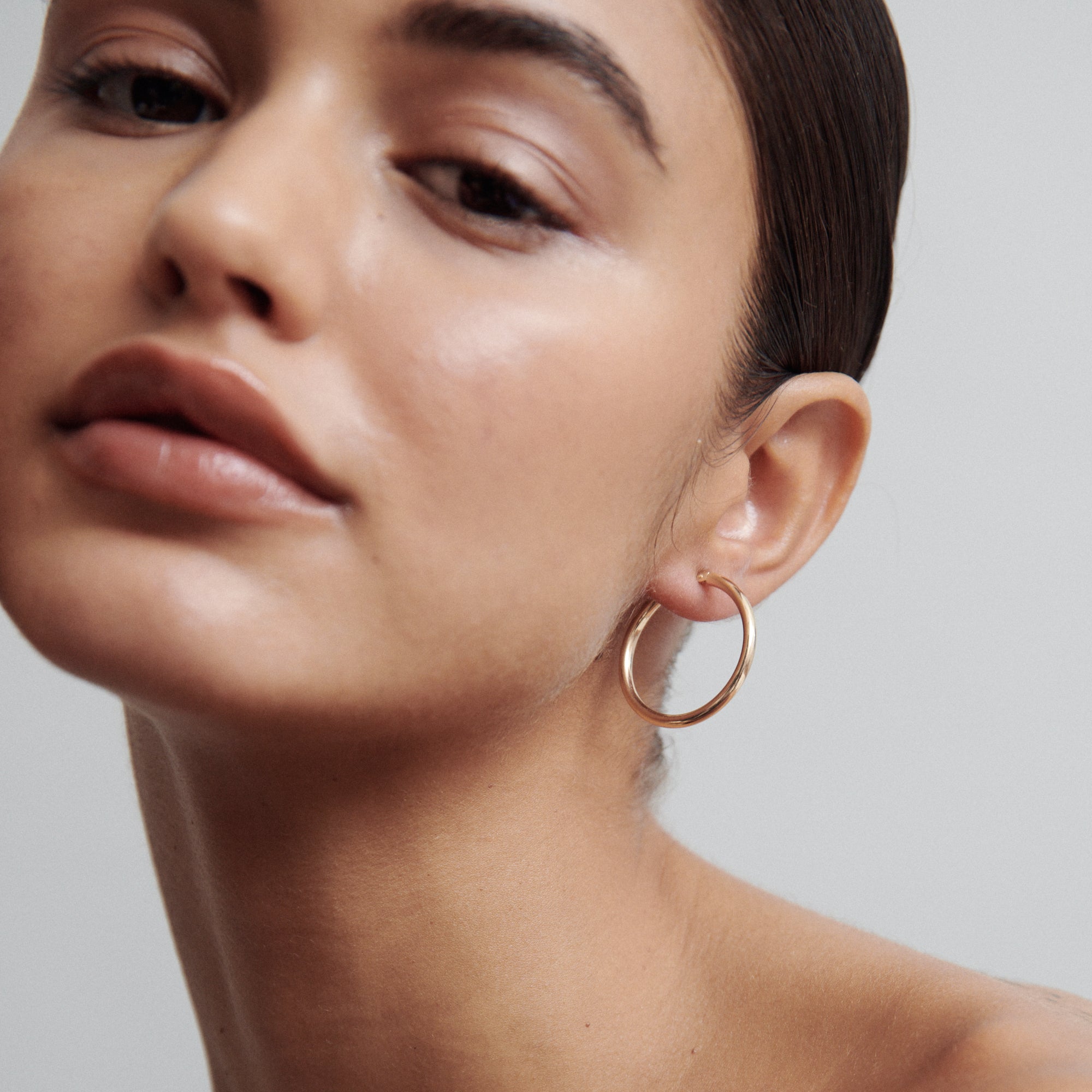 Close-up of a woman wearing a large gold hoop earring, showcasing luxury jewellery and flawless skin, with a neutral background.