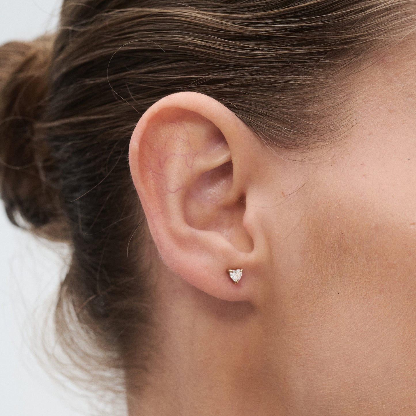 Close-up of an ear adorned with a small, elegant heart-shaped diamond stud earring. Hair is neatly tied back, highlighting the jewellery.