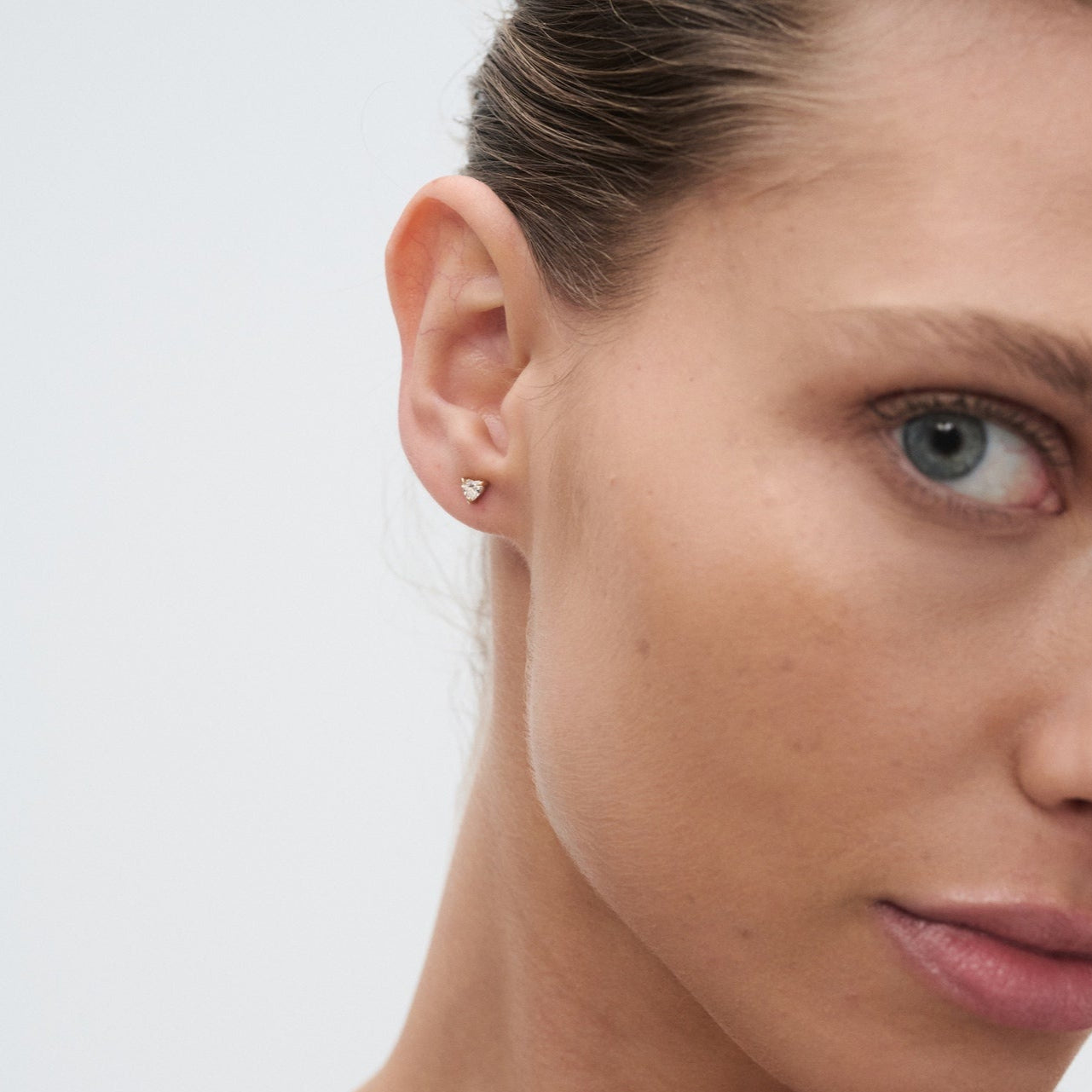 Close-up of a woman wearing a small, elegant diamond stud earring. Her hair is tied back, highlighting the jewellery against clear skin.