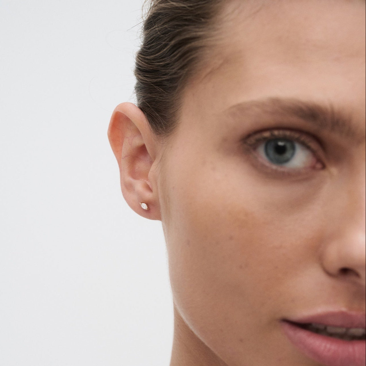 Close-up of a woman's face wearing a minimalist gold stud earring, highlighting luxury jewellery on smooth skin with a neutral expression.