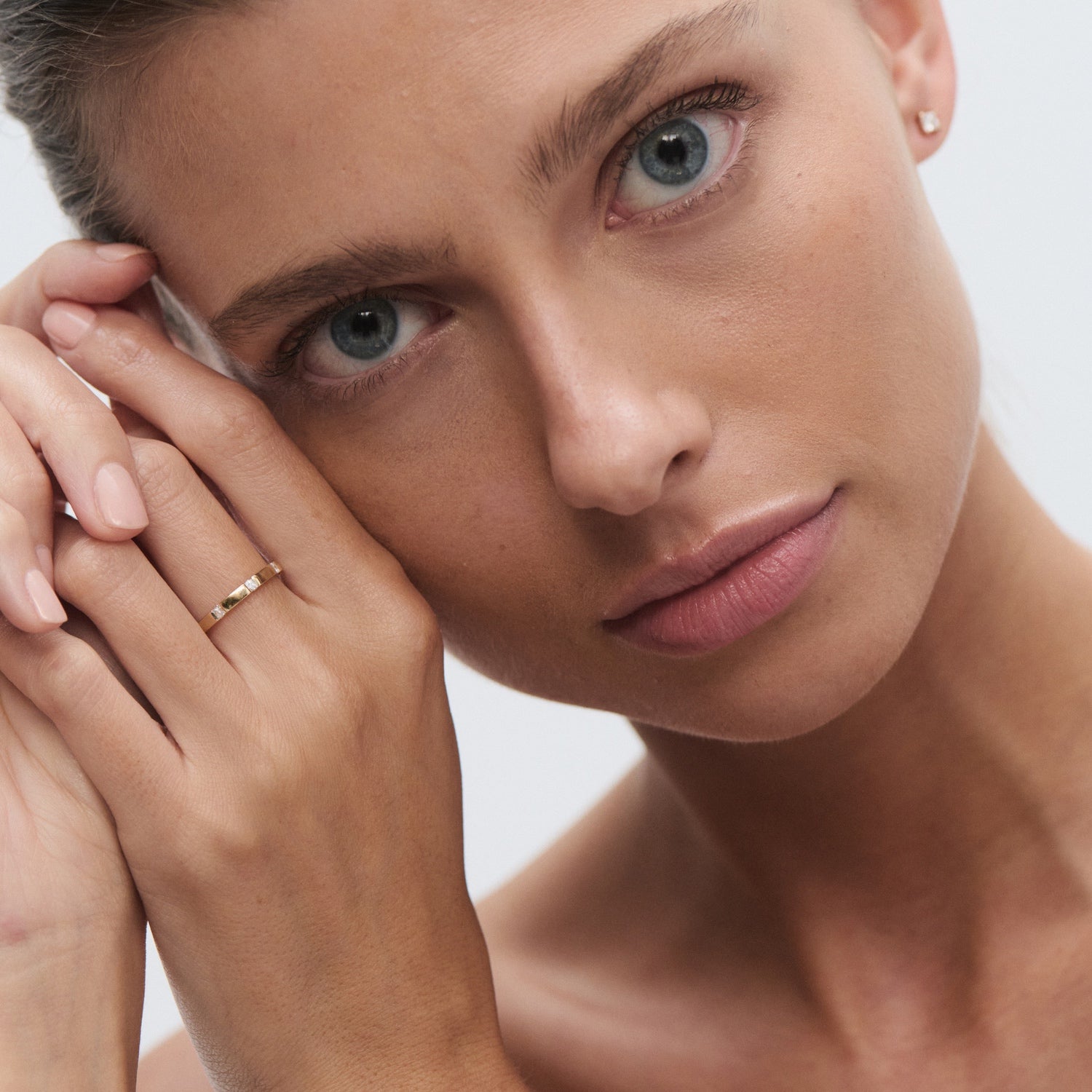 Close-up of a person with light eyes, wearing a gold ring and diamond stud earrings, gently touching their cheek with their hand.