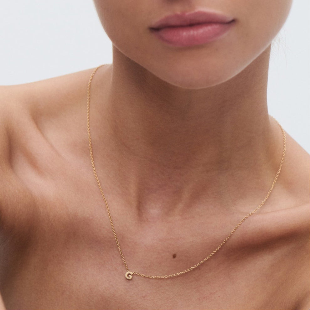 A woman wears a delicate gold necklace with a small pendant, set against a minimalistic background.