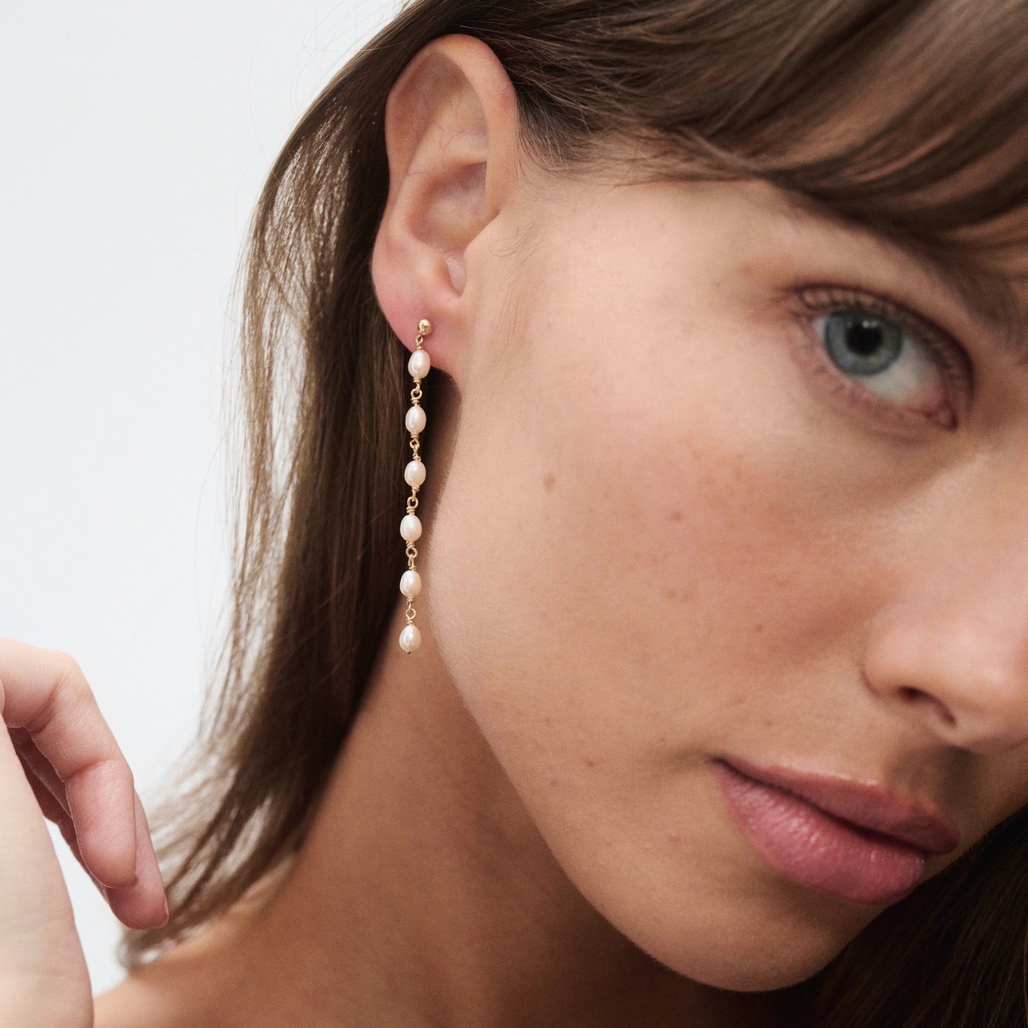 Woman wearing a long, elegant pearl drop earring on her left ear, with a light skin tone and natural makeup. Focus on jewellery details.
