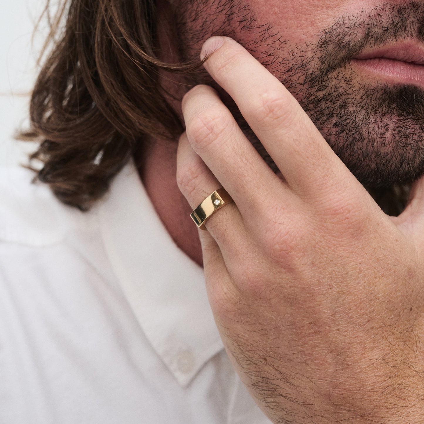 Man wearing a sophisticated gold ring with a small diamond, touching his cheek. He has a beard and long hair, wearing a white shirt.