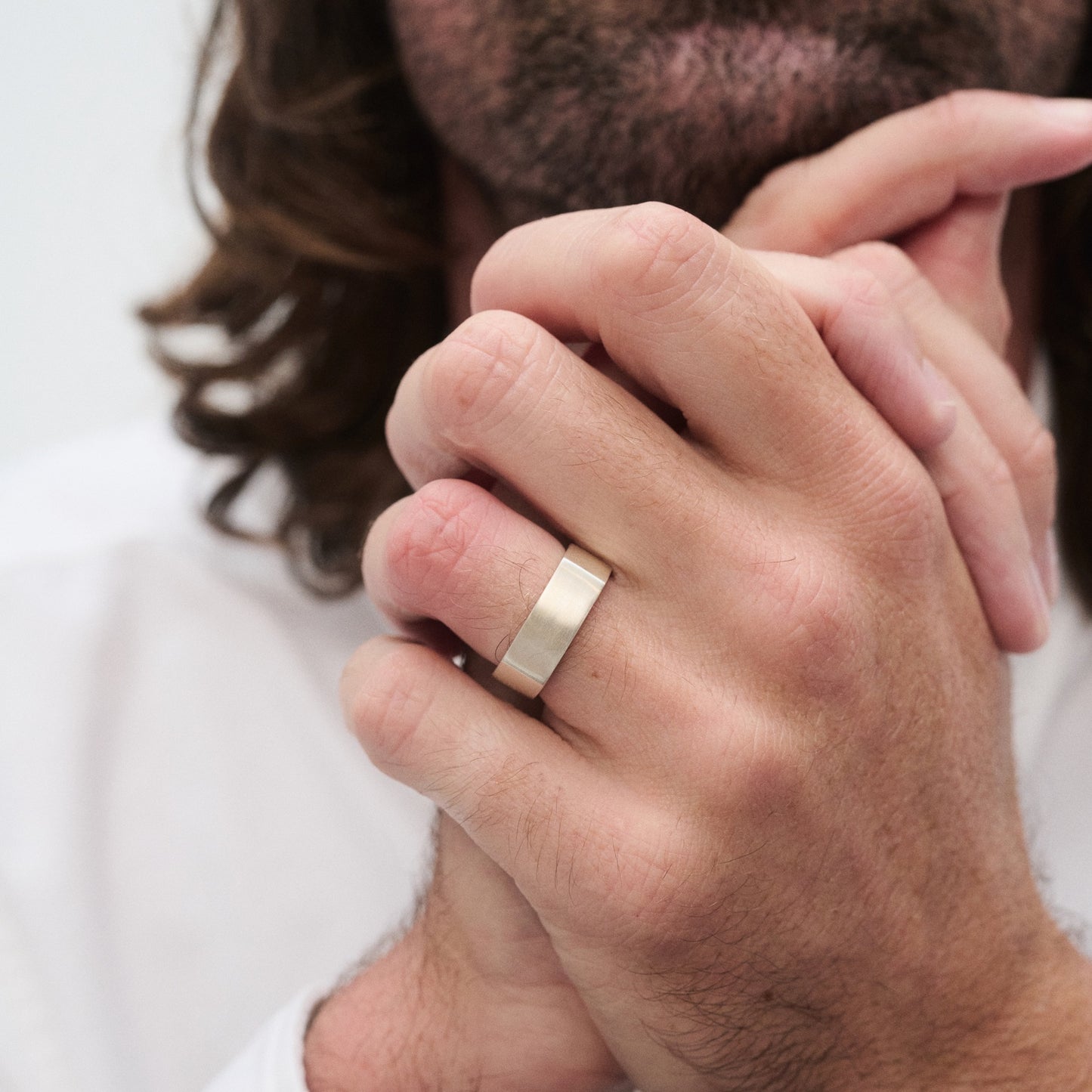 Man with long hair wearing a gold ring on his finger, hands clasped in front of a white shirt. Elegantly styled luxury jewellery focus.