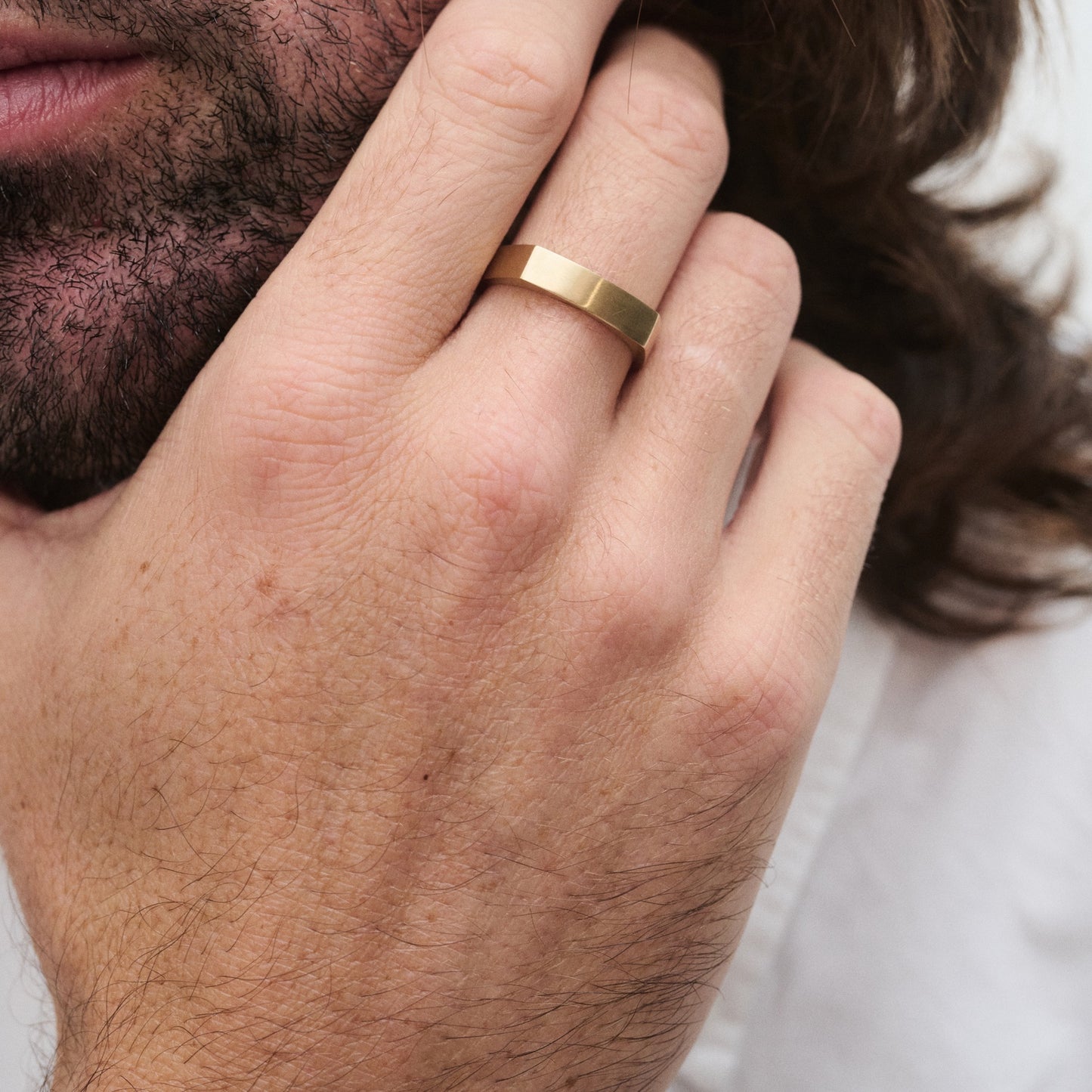 Close-up of a hand wearing a sleek gold ring on the index finger, with a textured beard and long hair in the background.