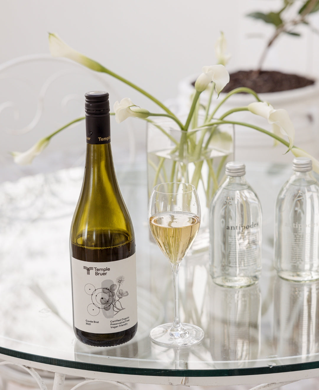 Wine bottle and glass on a clear glass table, accompanied by Antipodes water bottles and white flowers in background. Elegant setting.