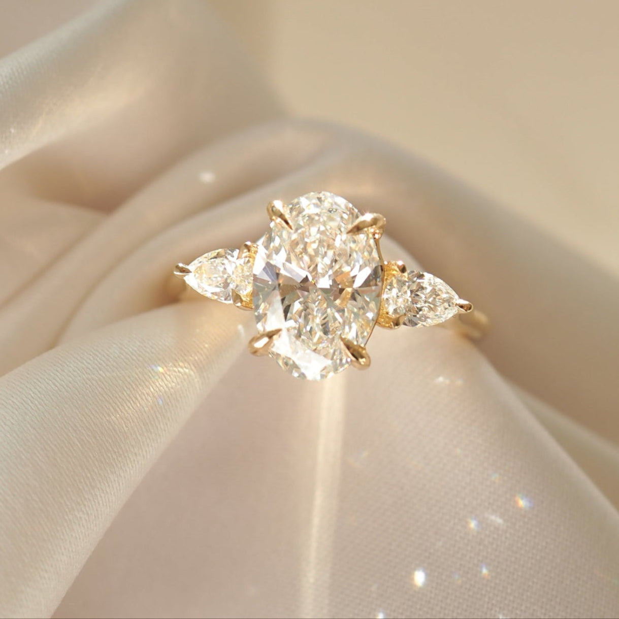 Elegant gold ring with a large oval diamond and two pear-shaped diamonds, resting on a silky fabric. Luxurious jewellery display.