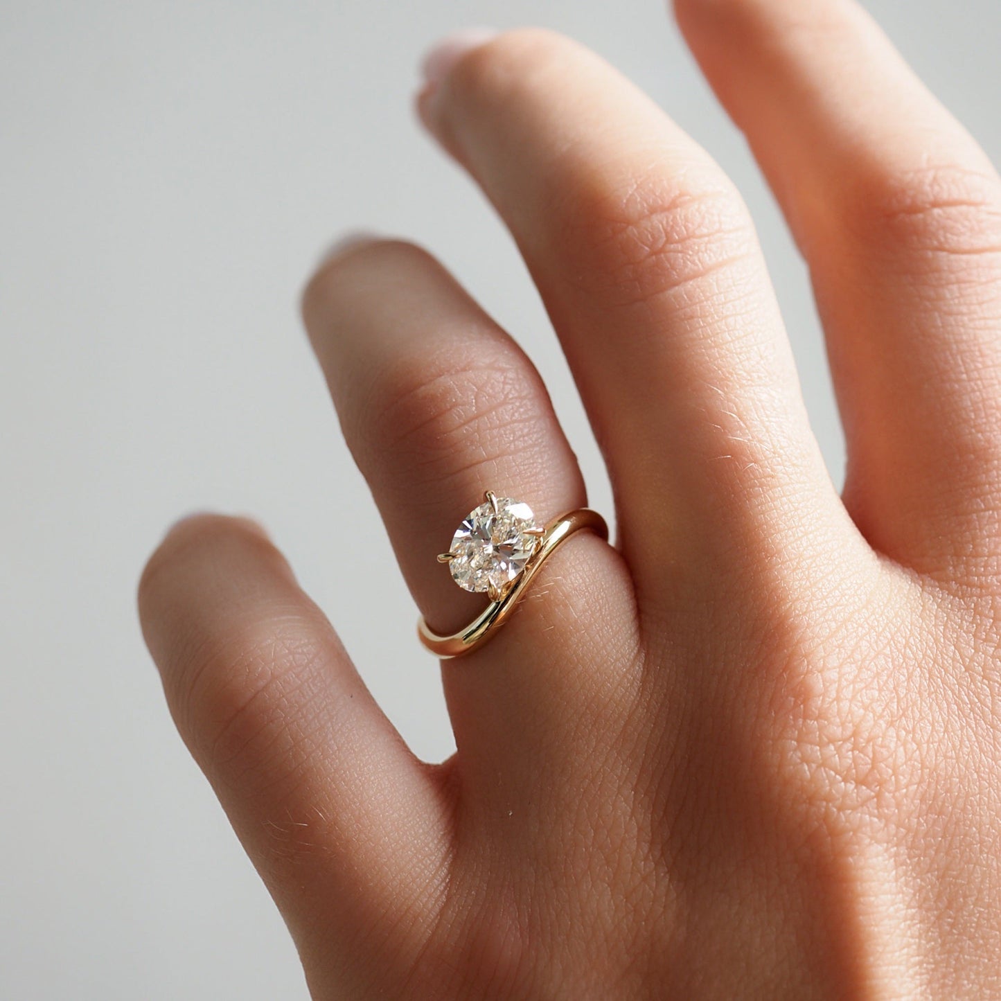 Hand wearing an elegant gold ring with a large, sparkling diamond solitaire, showcased against a soft grey background.