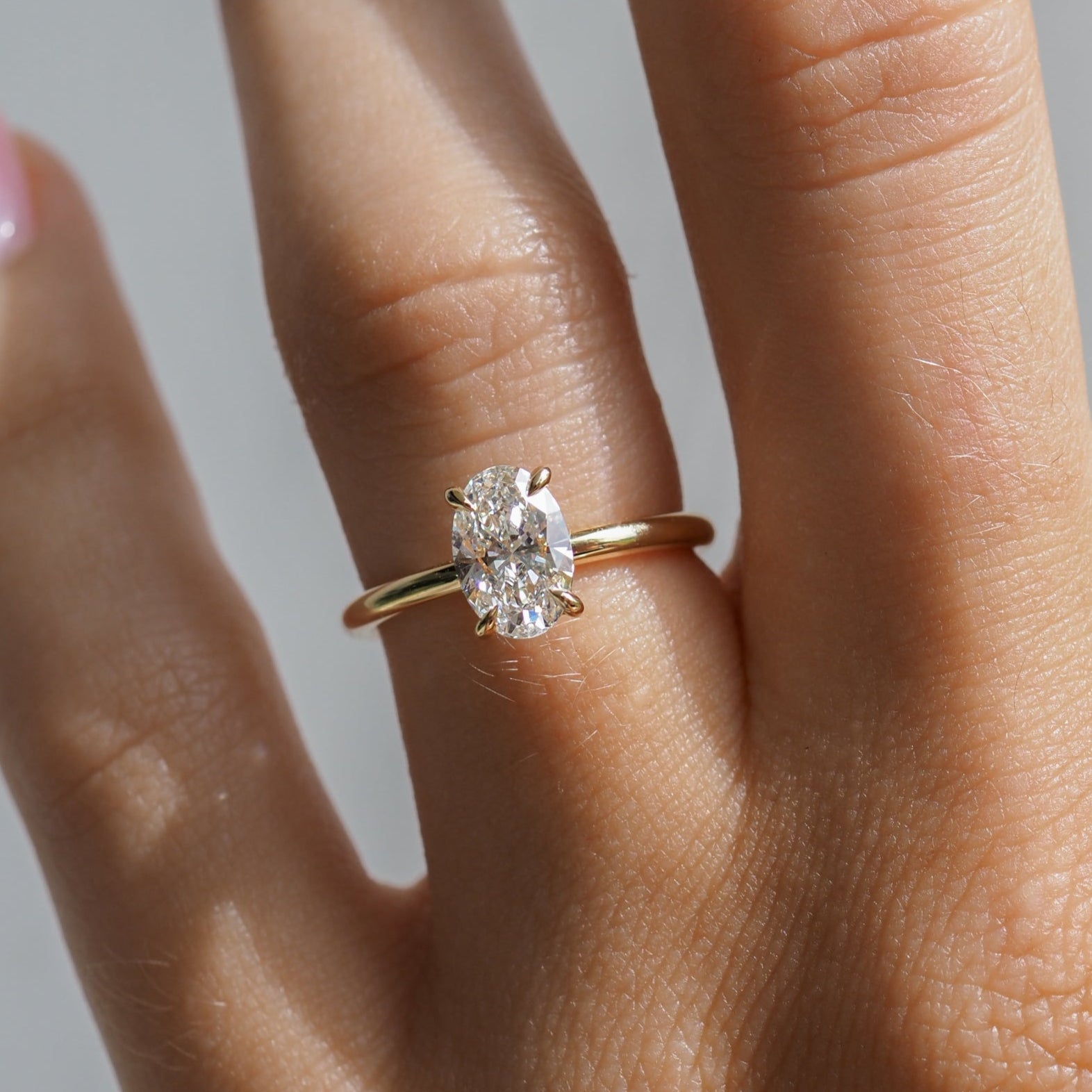 Hand with a gold ring featuring an oval diamond, sunlight highlighting the stone's brilliance. Finger with pastel pink nail polish.