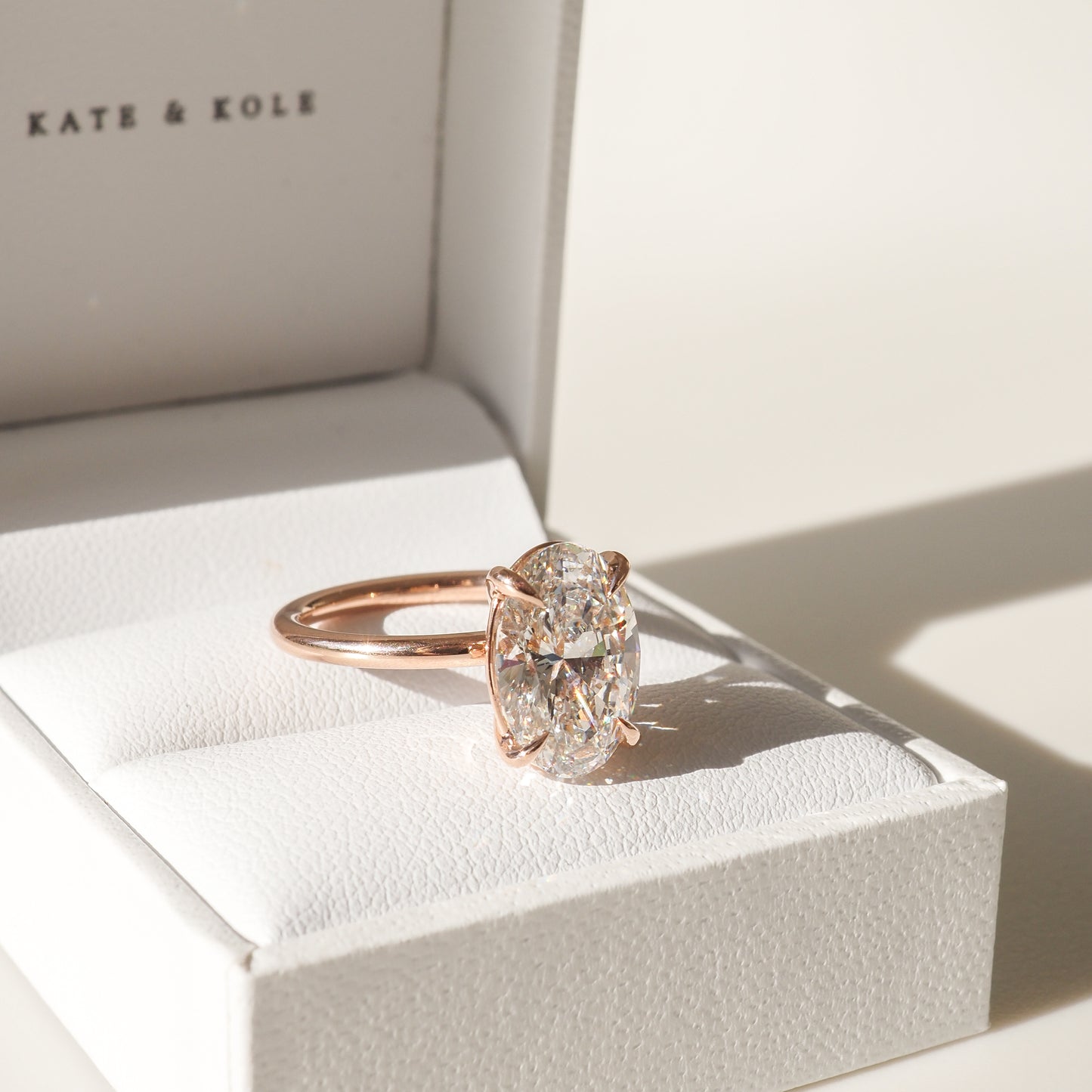 A solitaire diamond ring with a rose gold band on a white jewellery box, bathed in warm light.