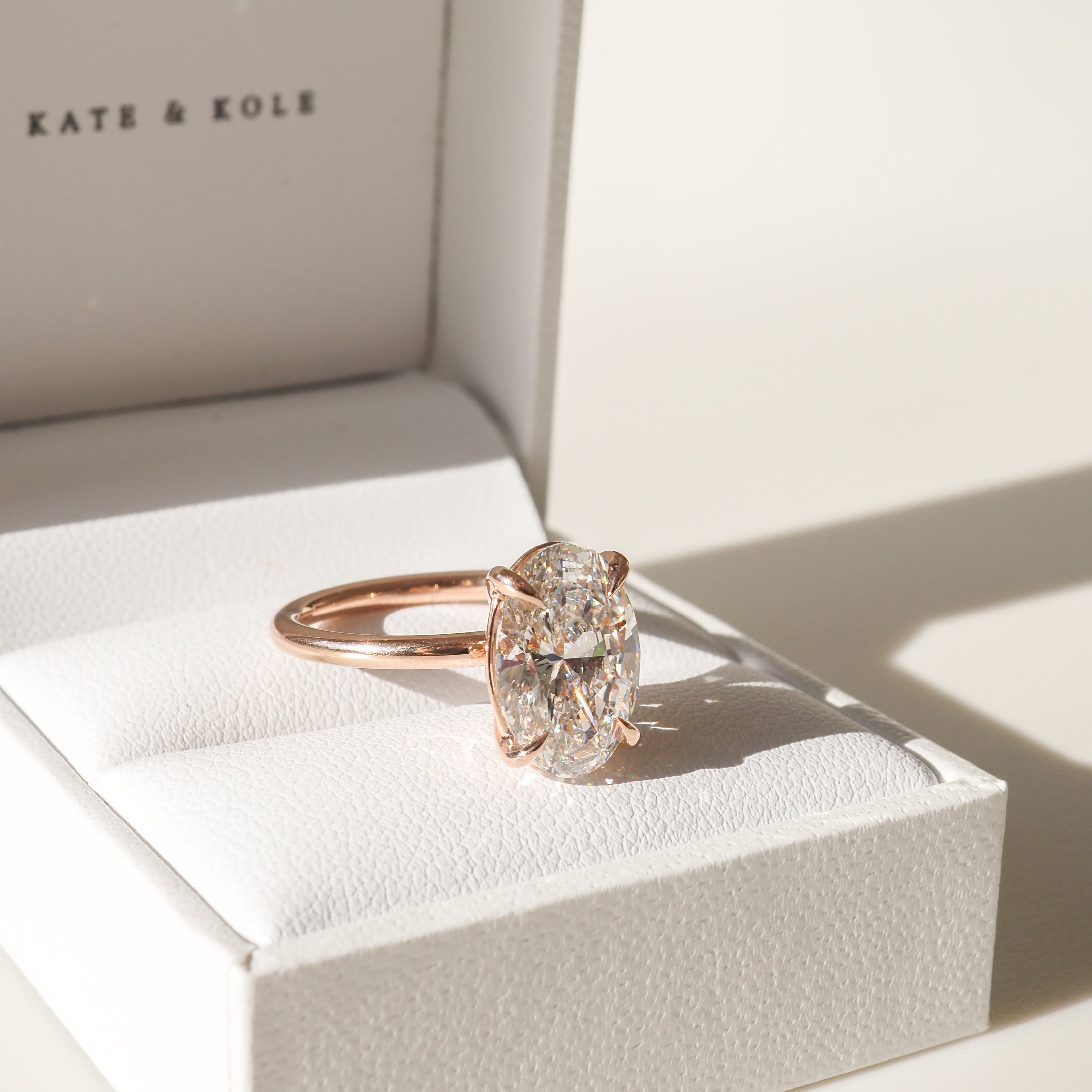 A solitaire diamond ring with a rose gold band on a white jewellery box, bathed in warm light.