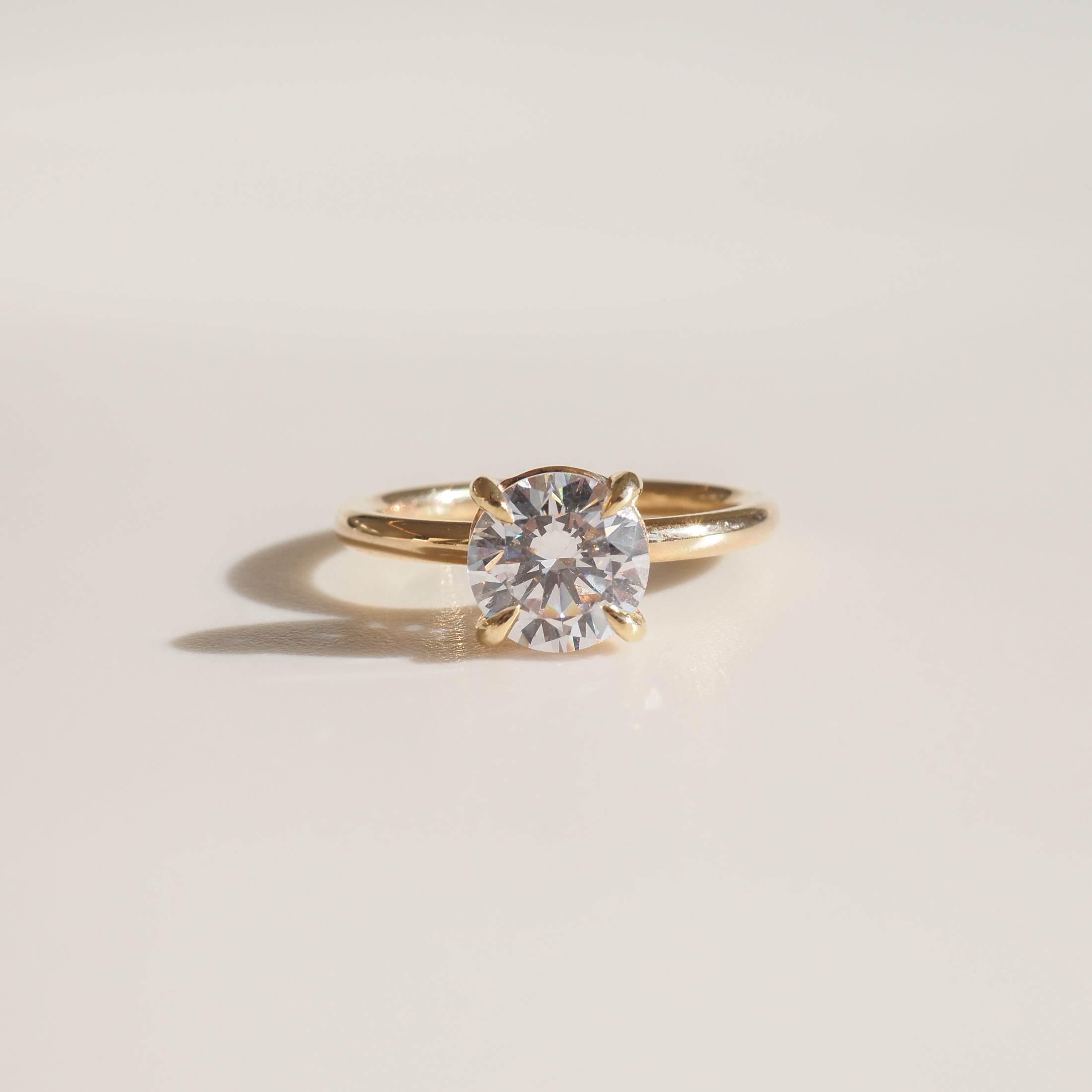 Gold solitaire engagement ring with a round brilliant diamond, elegantly set on a polished band, casting a soft shadow on a light surface.