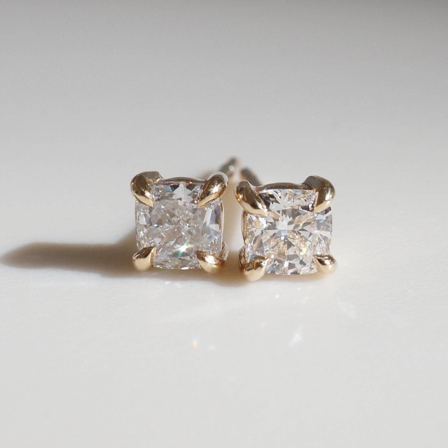 Close-up of cushion-cut diamond stud earrings set in gold, displayed on a reflective surface, highlighting luxury jewellery elegance.