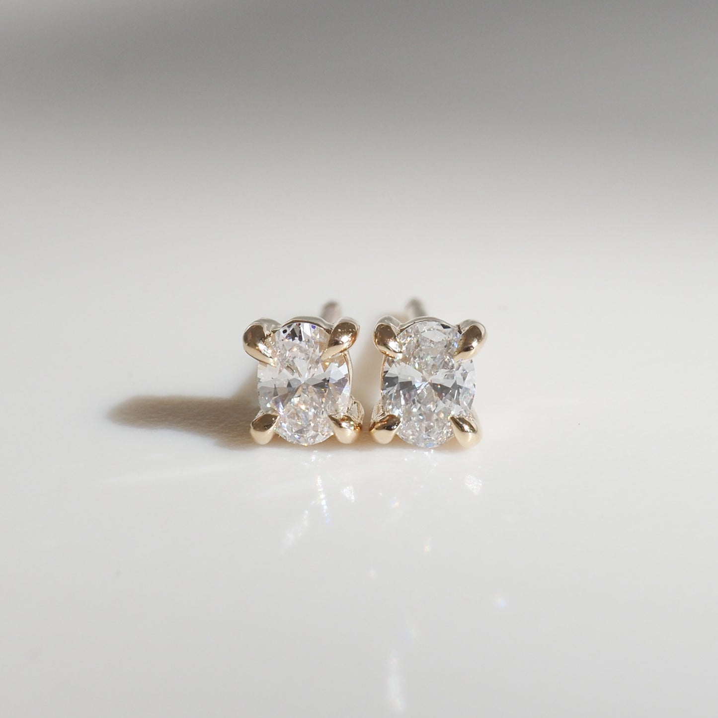 Pair of diamond stud earrings with gold prongs, elegantly showcased on a smooth, light grey surface, emitting a subtle, luxurious sparkle.