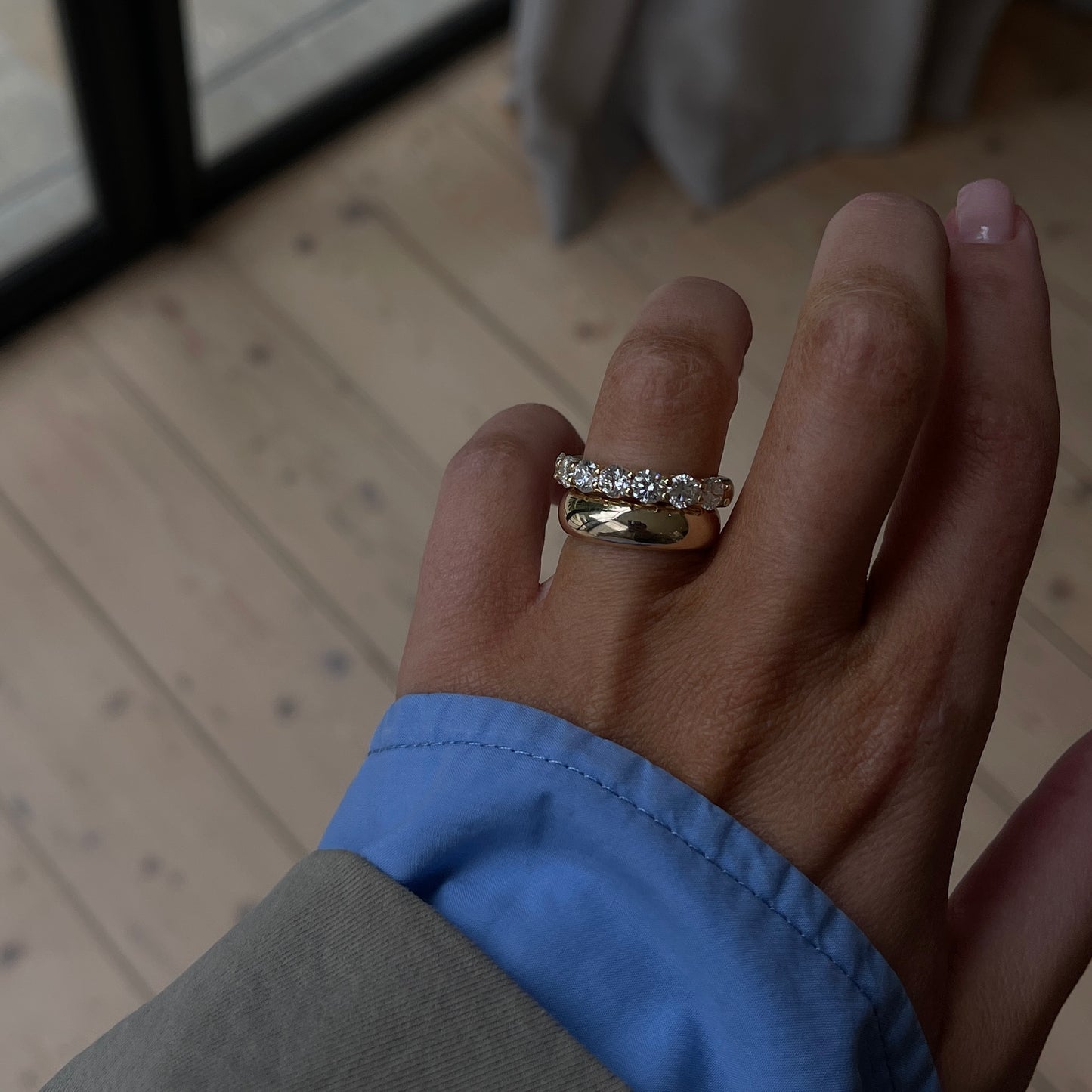 Hand wearing two gold rings, one plain and one set with diamonds, against a light wooden floor background. Elegant, luxury jewellery.
