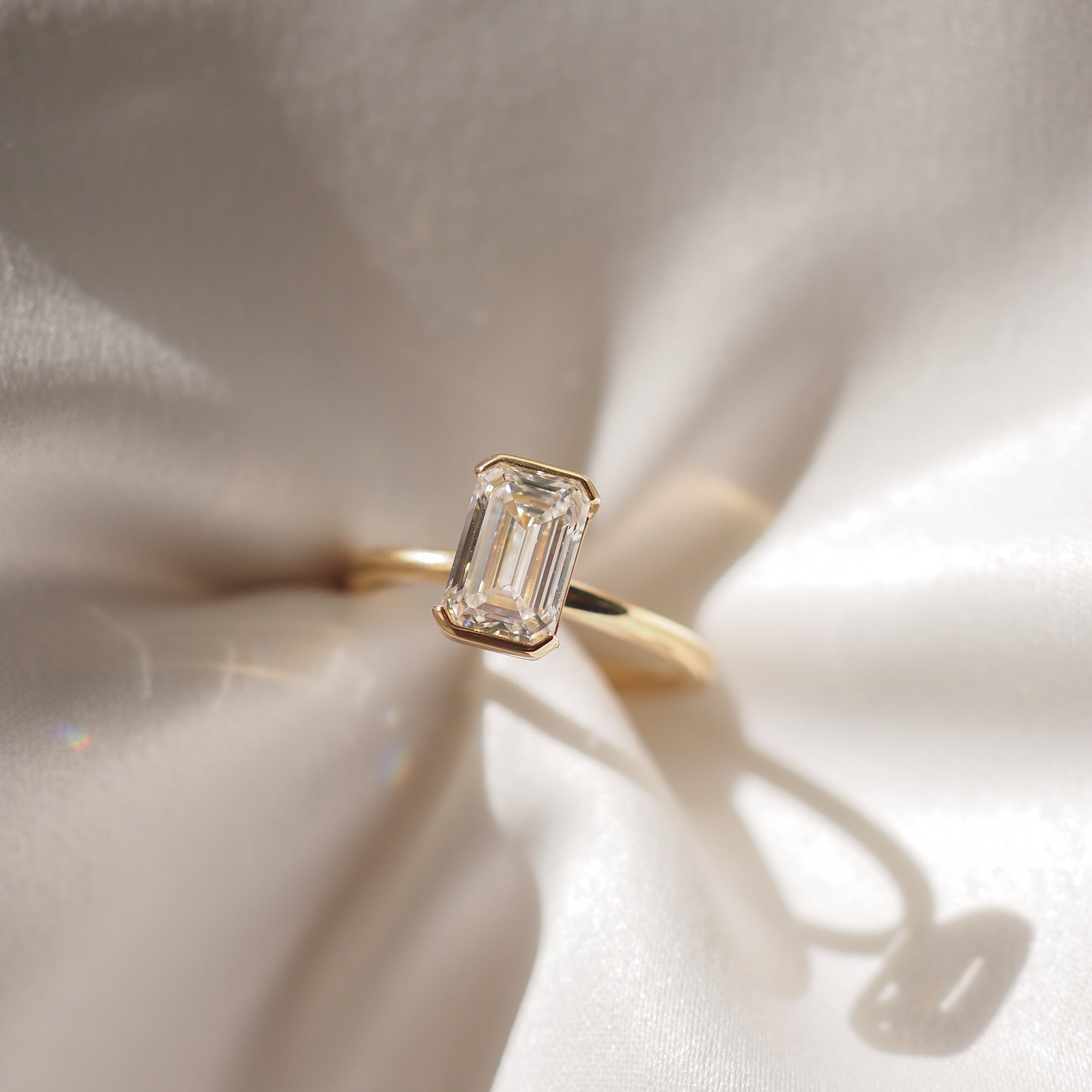 Emerald-cut diamond ring set on gold band, elegantly displayed on smooth, sunlit silk fabric, showcasing luxury jewellery craftsmanship.