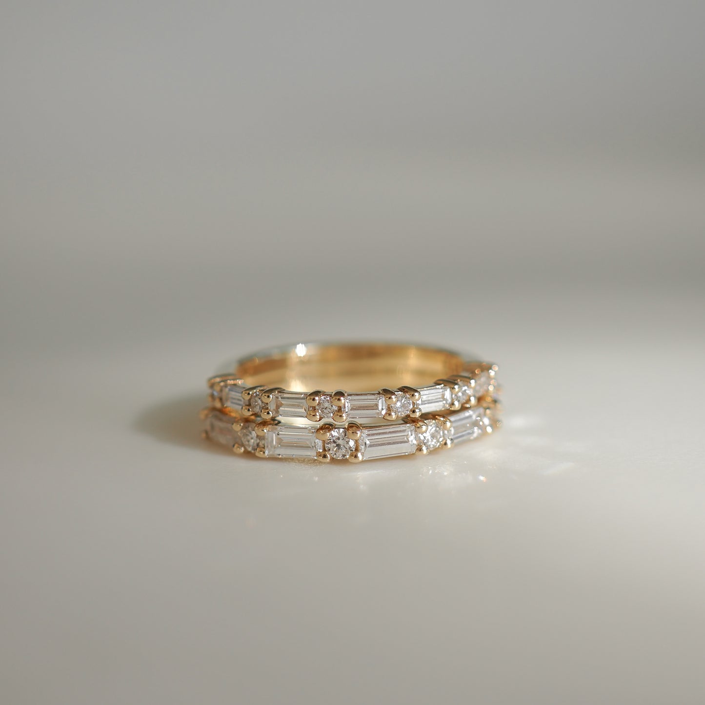 Gold ring featuring two rows of radiant diamond baguettes, elegantly stacked, set against a soft grey background.