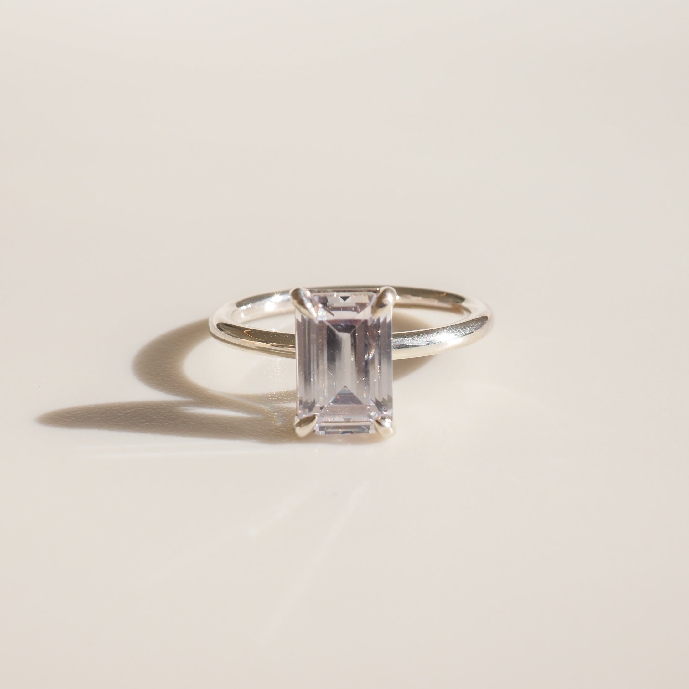 Elegant silver ring with a rectangular-cut clear gemstone, casting a shadow on a light surface; luxury jewellery photography.