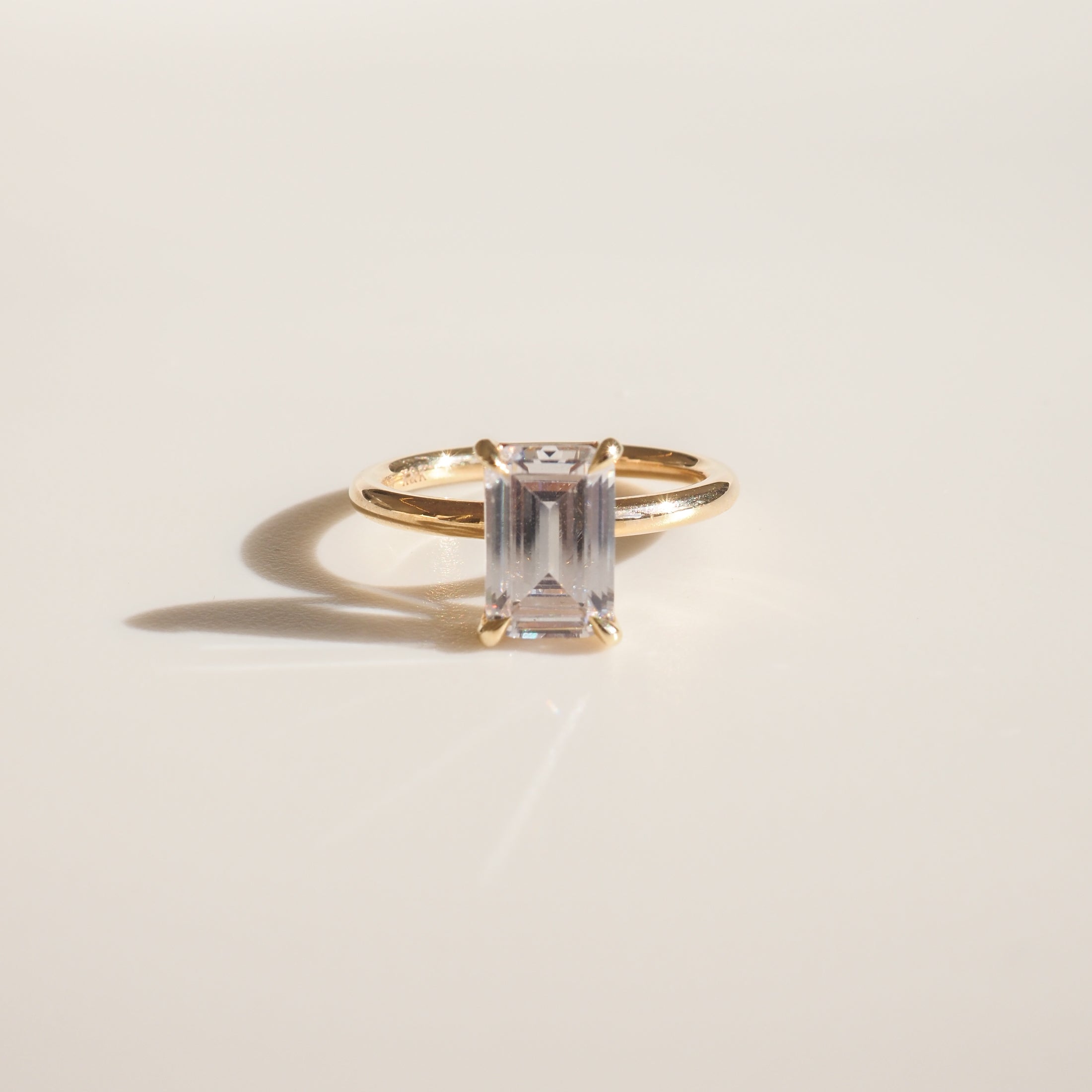Gold ring with an emerald-cut clear gemstone on a smooth cream background, casting a delicate shadow. Perfect for luxury jewellery enthusiasts.