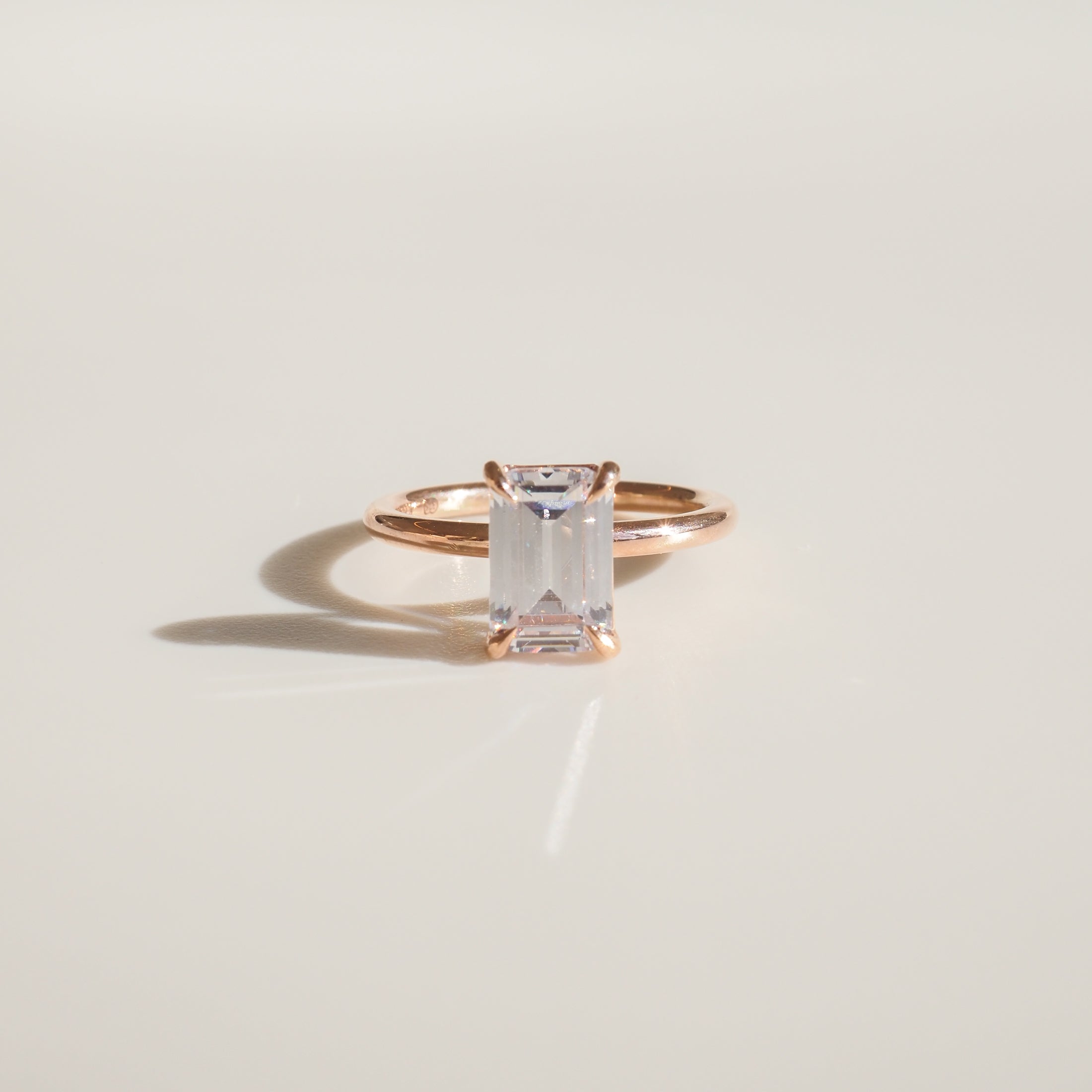 Emerald-cut diamond ring in rose gold setting on a soft beige background, casting a subtle shadow. Luxurious minimalist jewellery design.