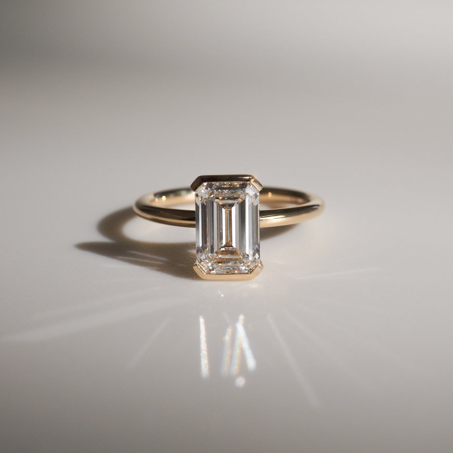 Emerald-cut diamond ring with a gold band, elegantly reflecting light on a smooth surface, highlighting luxury jewellery design.