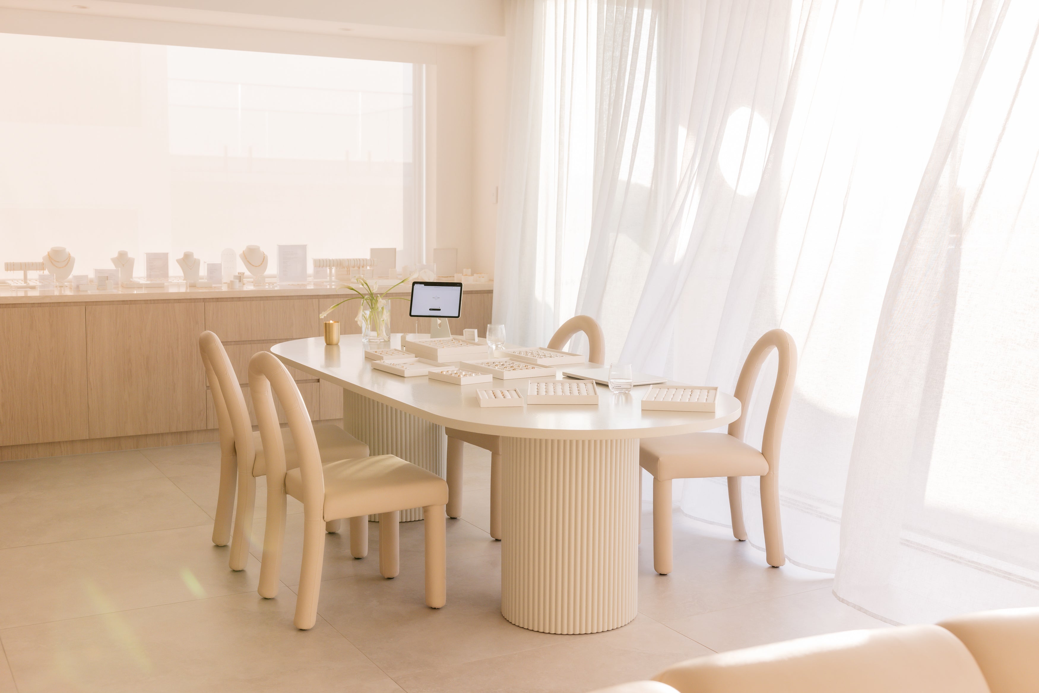 Luxurious jewellery showroom with elegant cream furniture, large table displaying jewellery boxes, soft sunlight filtering through white curtains.