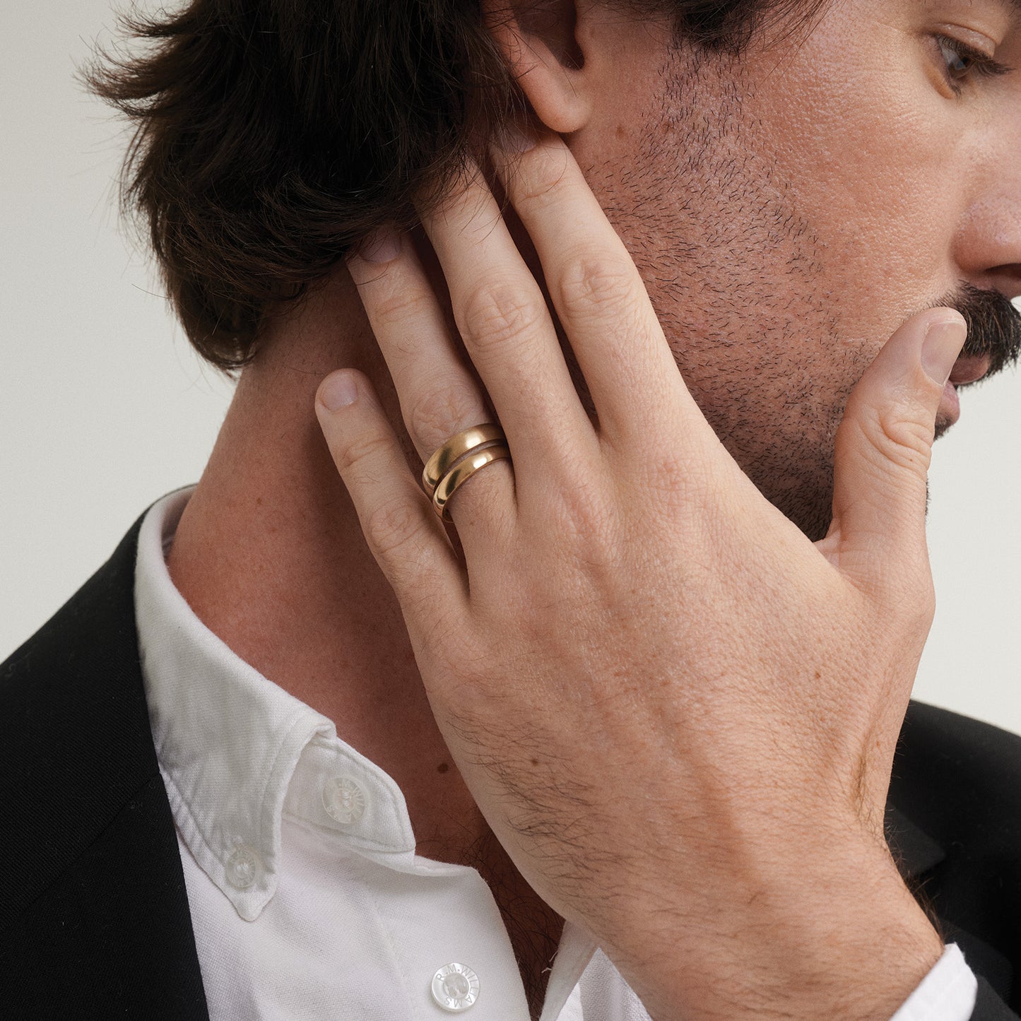 Man wearing two sizes of a gold ring on his hand, touching his ear against a neutral background