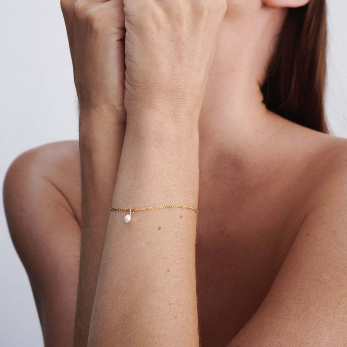 Woman wearing a delicate gold bracelet with a single white pearl, resting on her wrist, against a plain background.