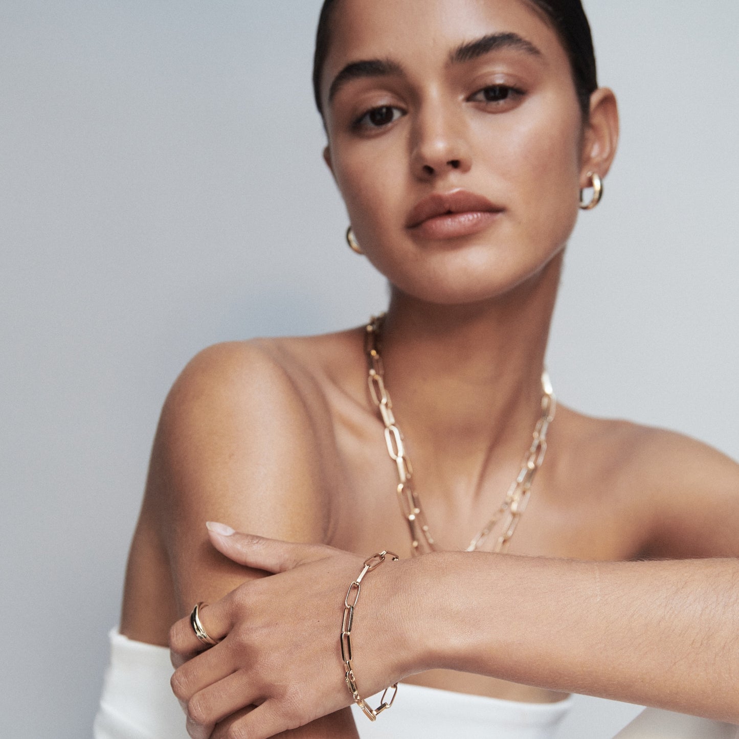 Woman wearing gold hoop earrings, layered chain necklaces, bracelet, and ring, against a neutral background. Luxe jewellery fashion.