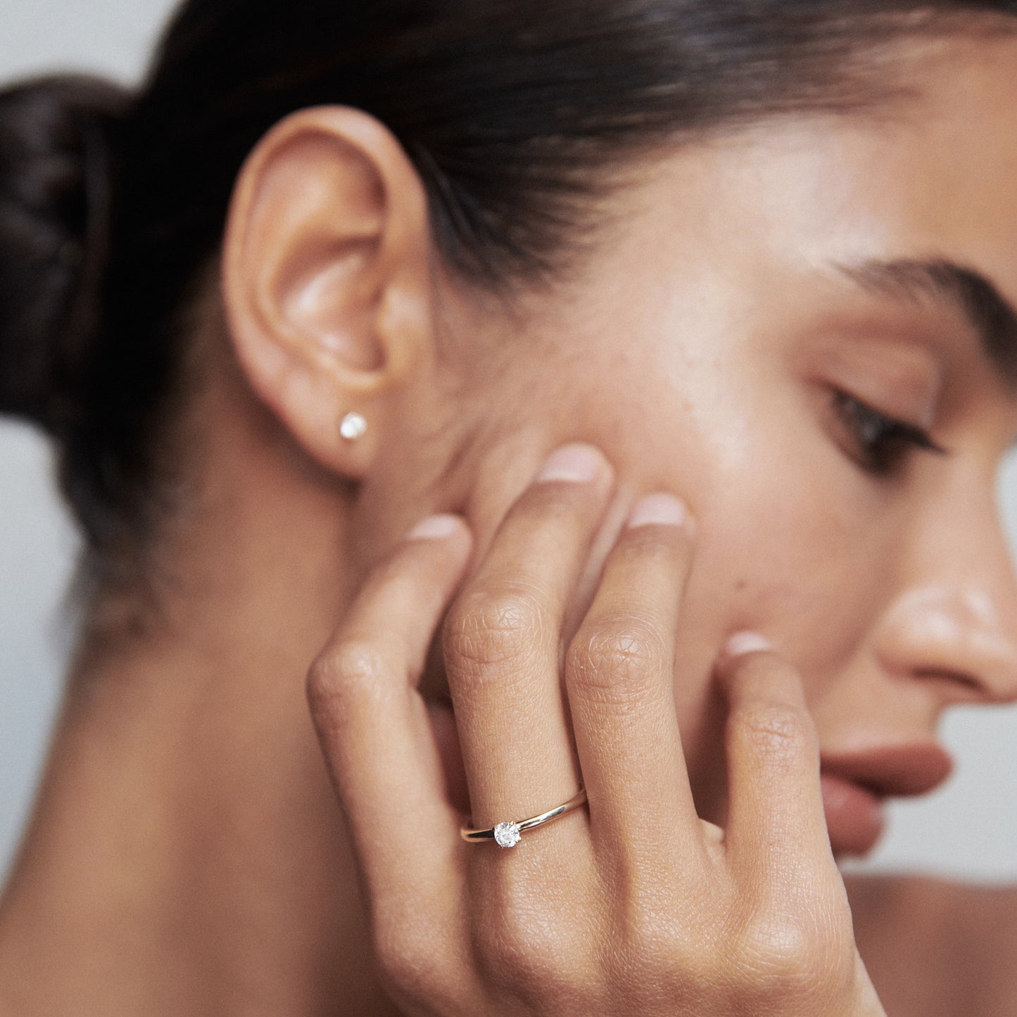 Woman wearing a diamond solitaire ring and small diamond stud earring, with a sleek hairstyle, showcasing luxury jewellery style.
