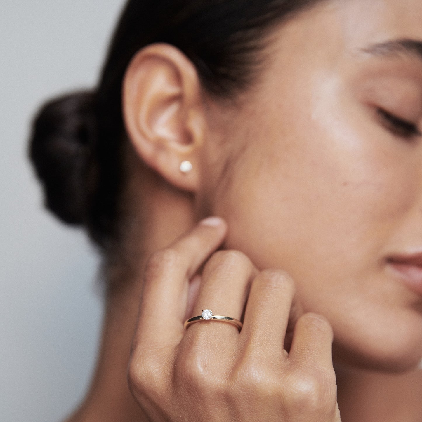 Woman wears a gold solitaire diamond ring and matching stud earring, eyes closed. Luxury jewellery, minimalist elegance.