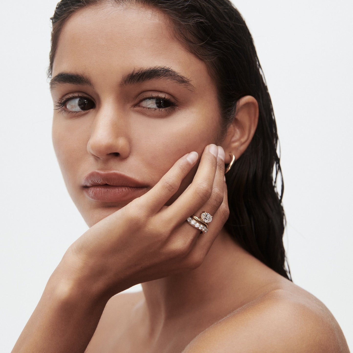 Woman with wet hair wearing gold hoop earrings and a stack of elegant gold rings adorned with diamonds. She gazes thoughtfully to the side.