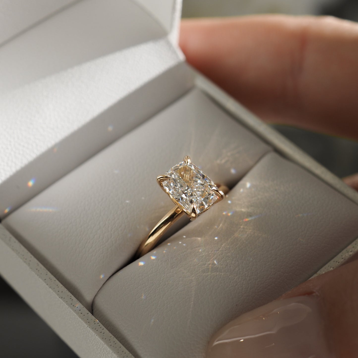 Gold engagement ring with radiant-cut diamond in a white jewellery box. Person holding the box, light reflecting off the diamond.