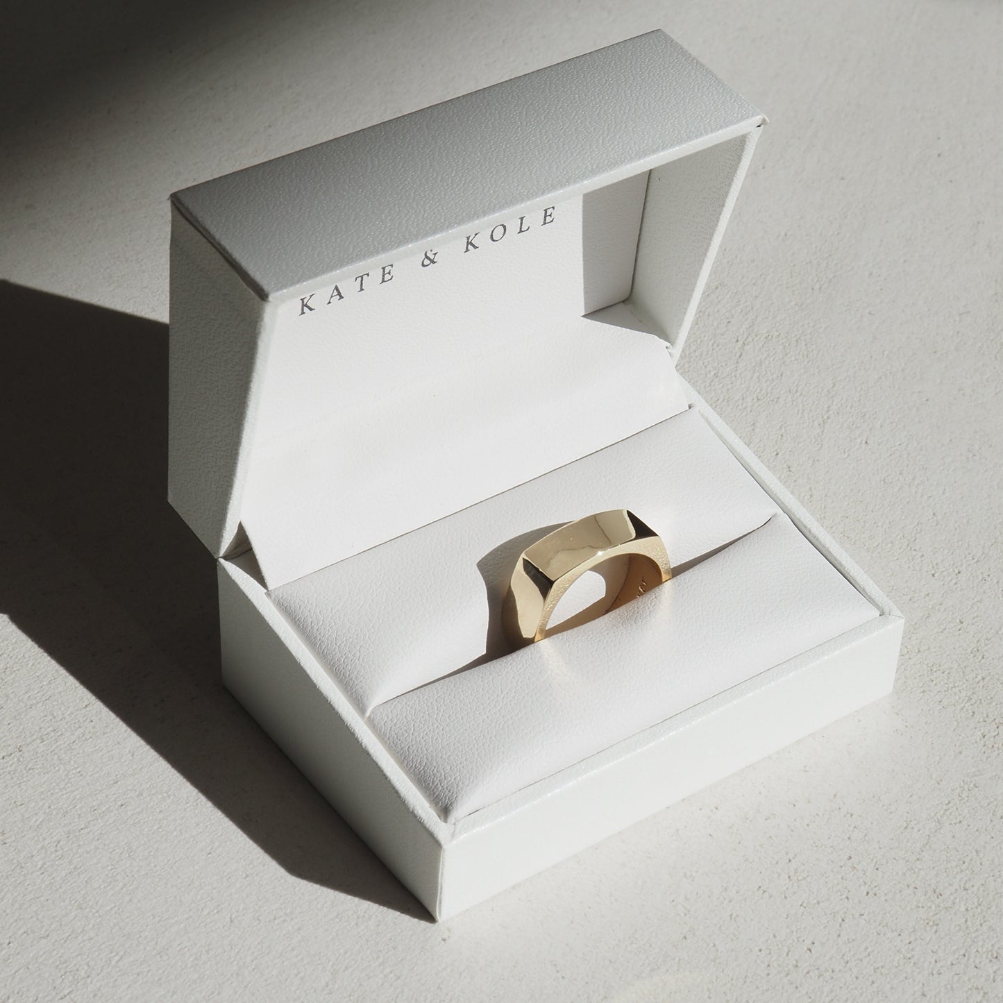 Gold geometric ring in a white Kate & Kole jewellery box, resting on a textured surface, highlighted by soft natural light.