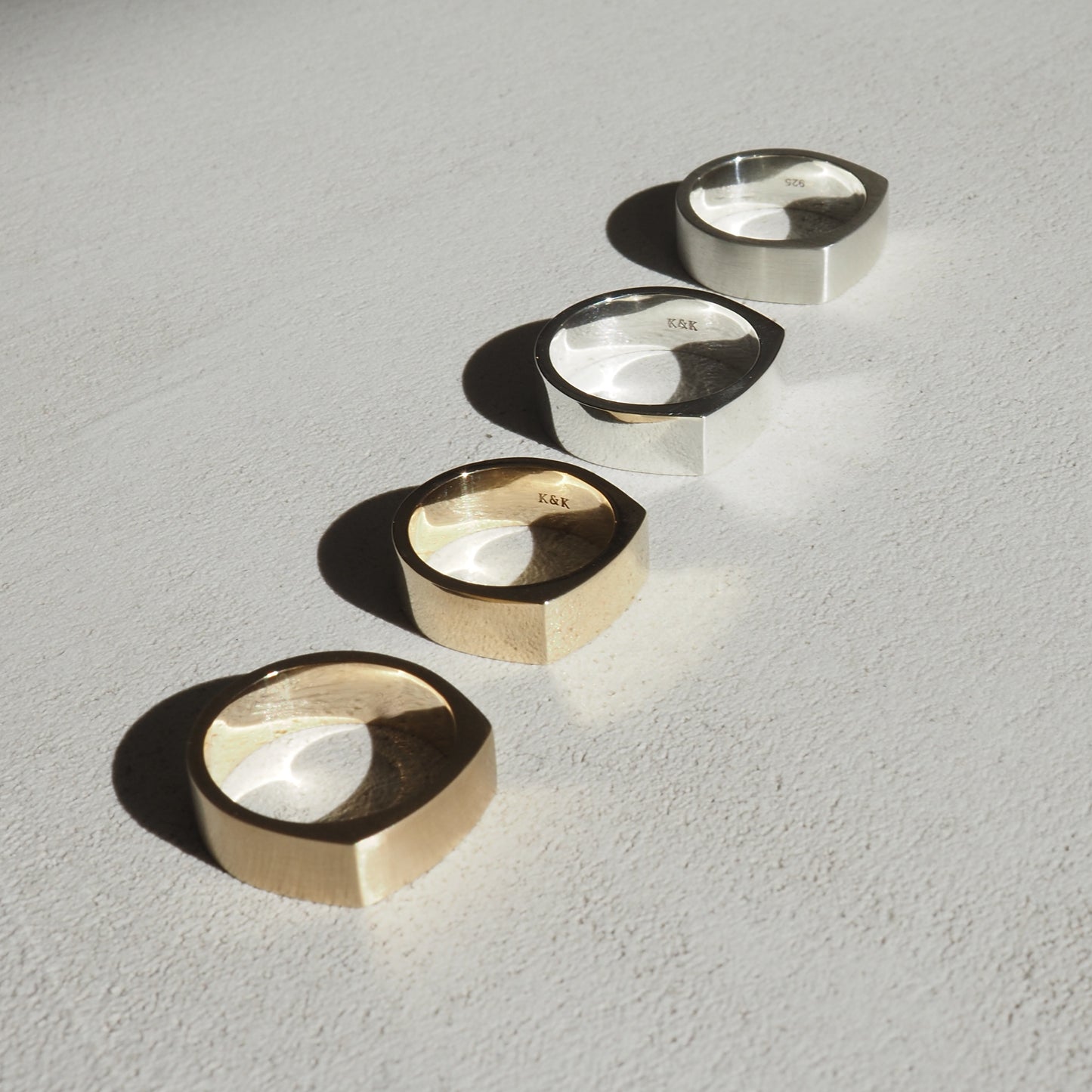 Four minimalist signet rings in gold and silver, artistically arranged on a textured white surface, casting soft shadows.