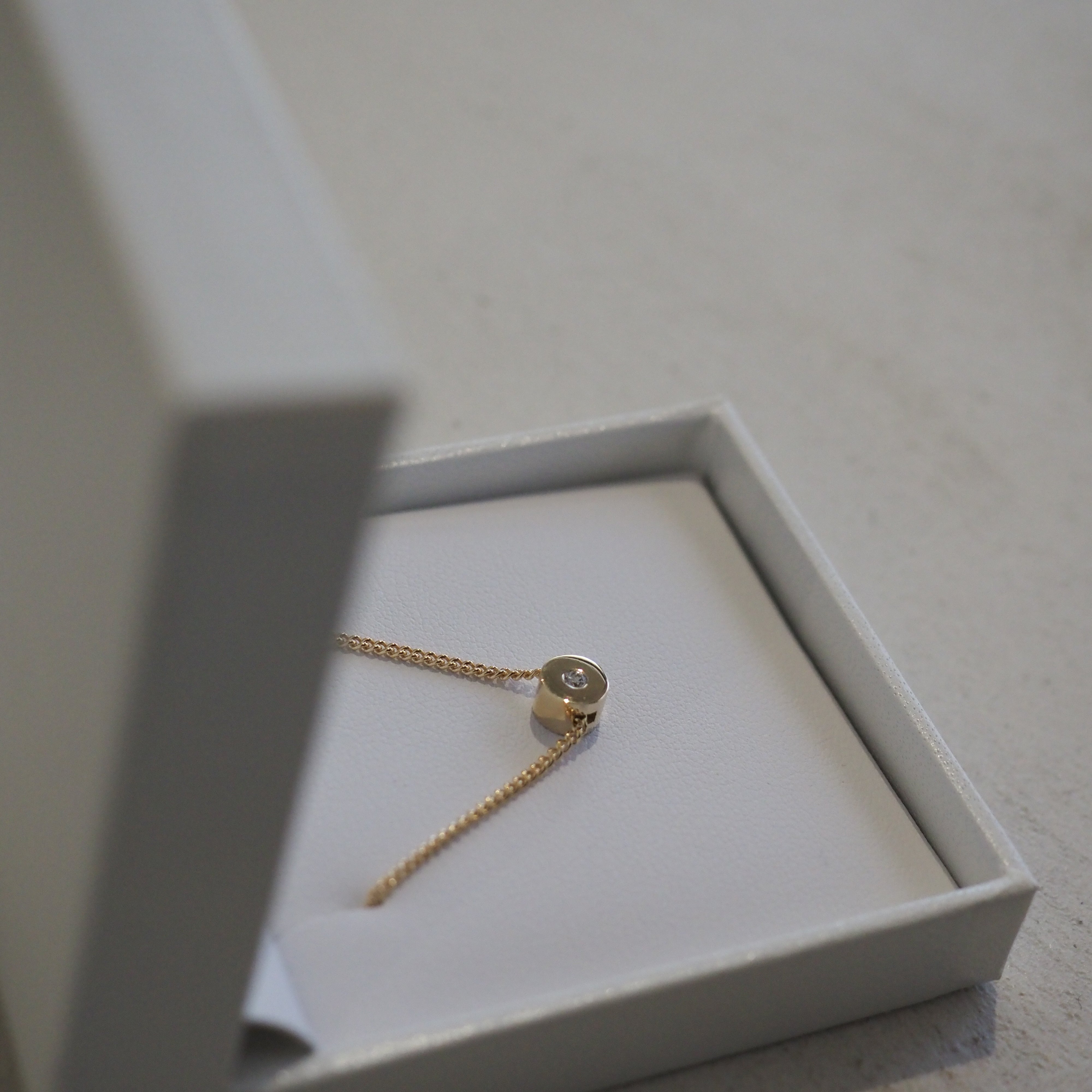 Gold chain necklace with a circular diamond pendant inside an open white jewellery box on a grey surface.