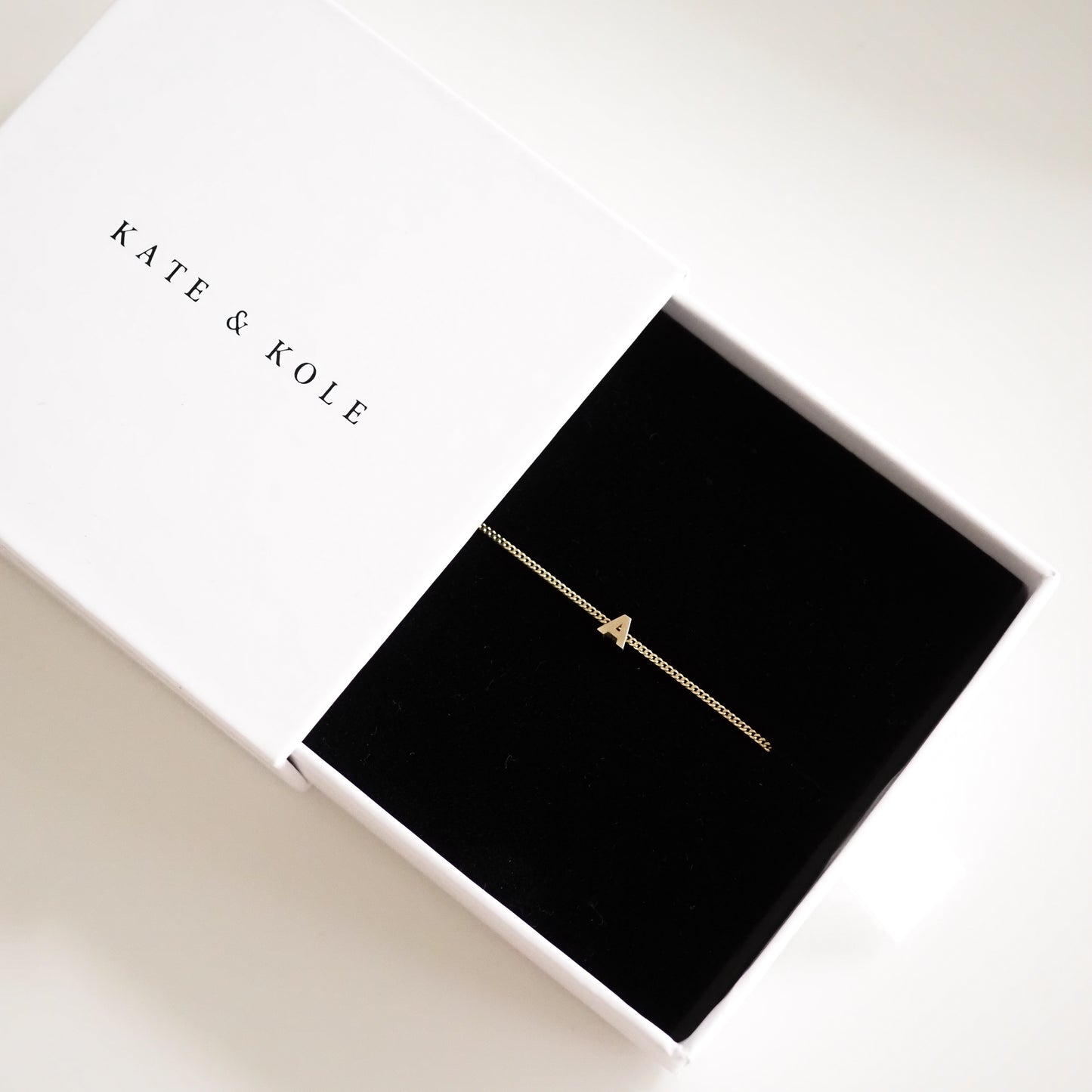 Gold bracelet with letter "A" on black velvet in a white Kate & Kole box; minimalist luxury jewellery design.