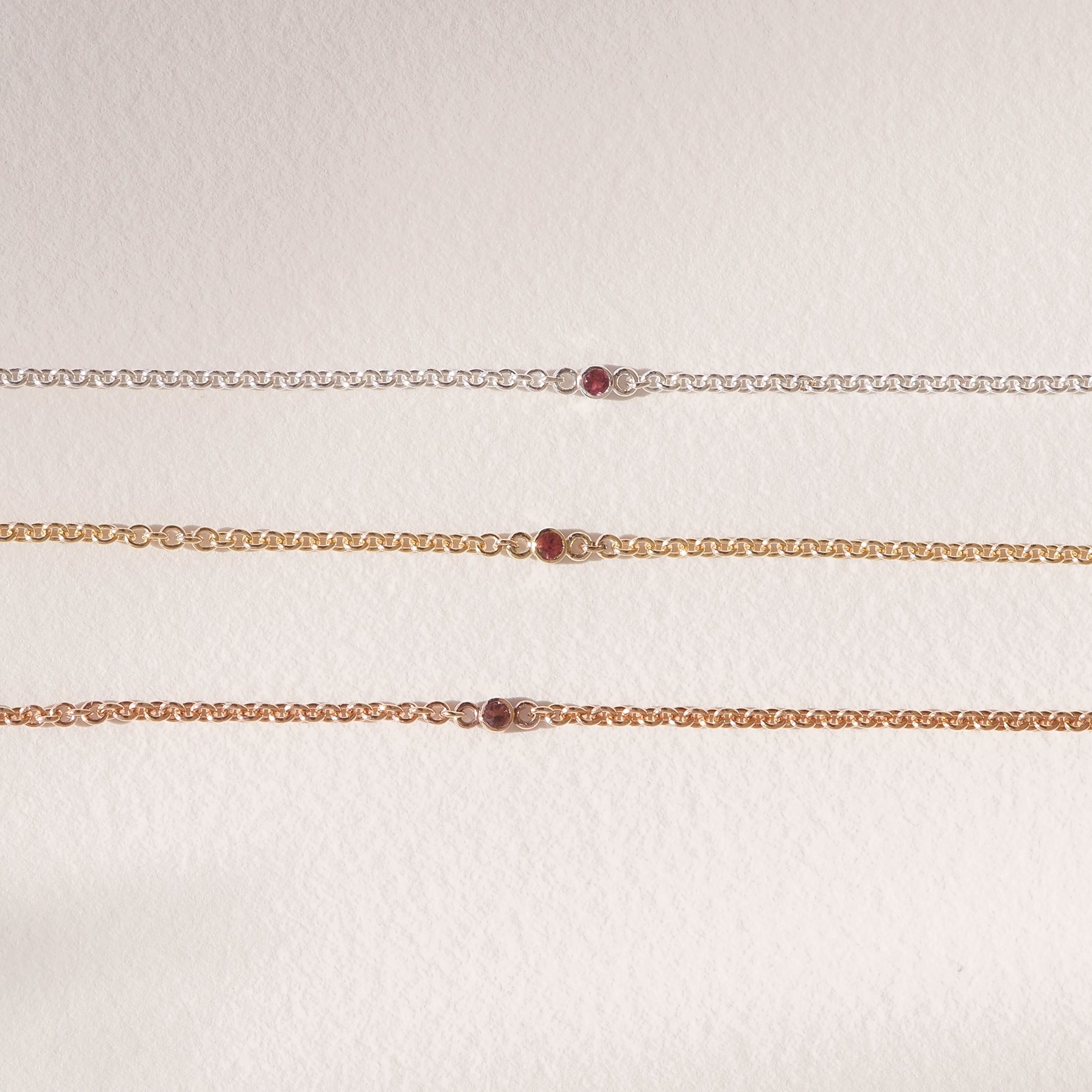 Three delicate chains in gold and silver display small red gemstones, elegantly laid out on a soft, cream background.