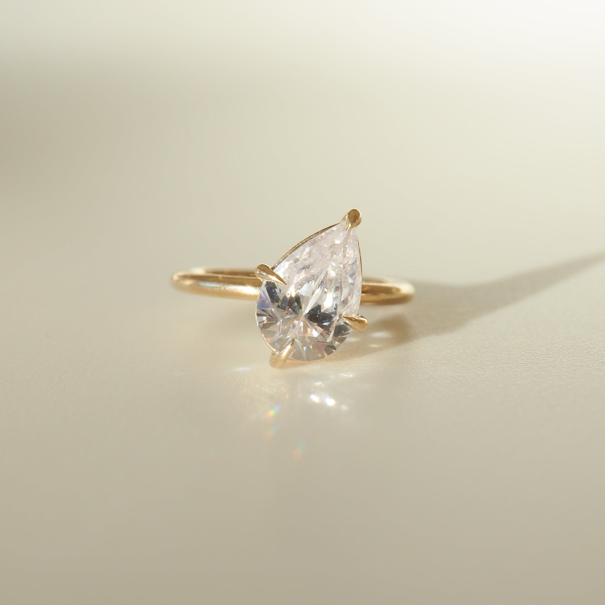 Gold ring with a pear-shaped diamond elegantly rests on a light surface, casting a soft shadow. Perfect for luxury jewellery lovers.