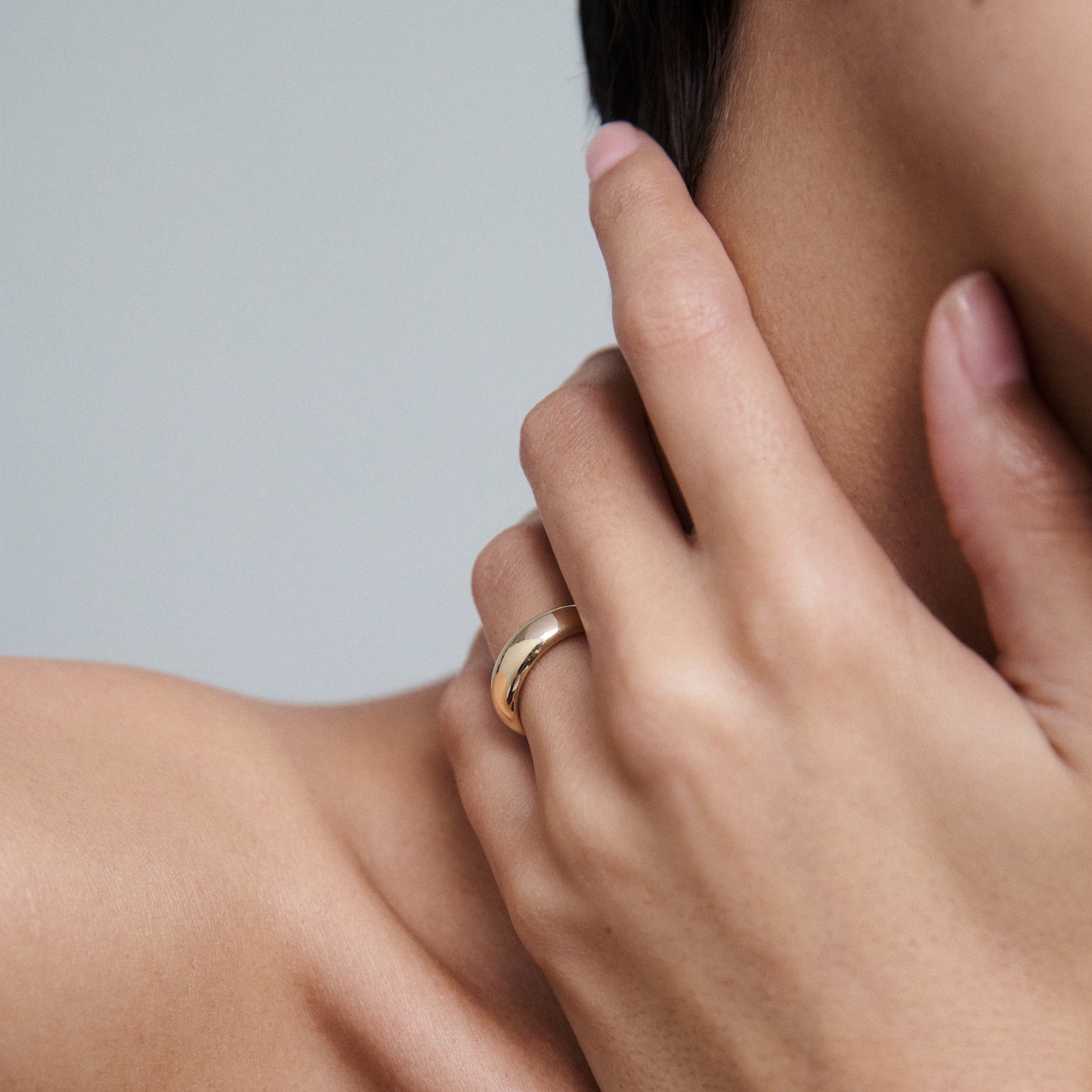 Close-up of a gold ring on a woman's hand near her neck, showcasing luxury jewellery. Minimalist and elegant design against a neutral background.