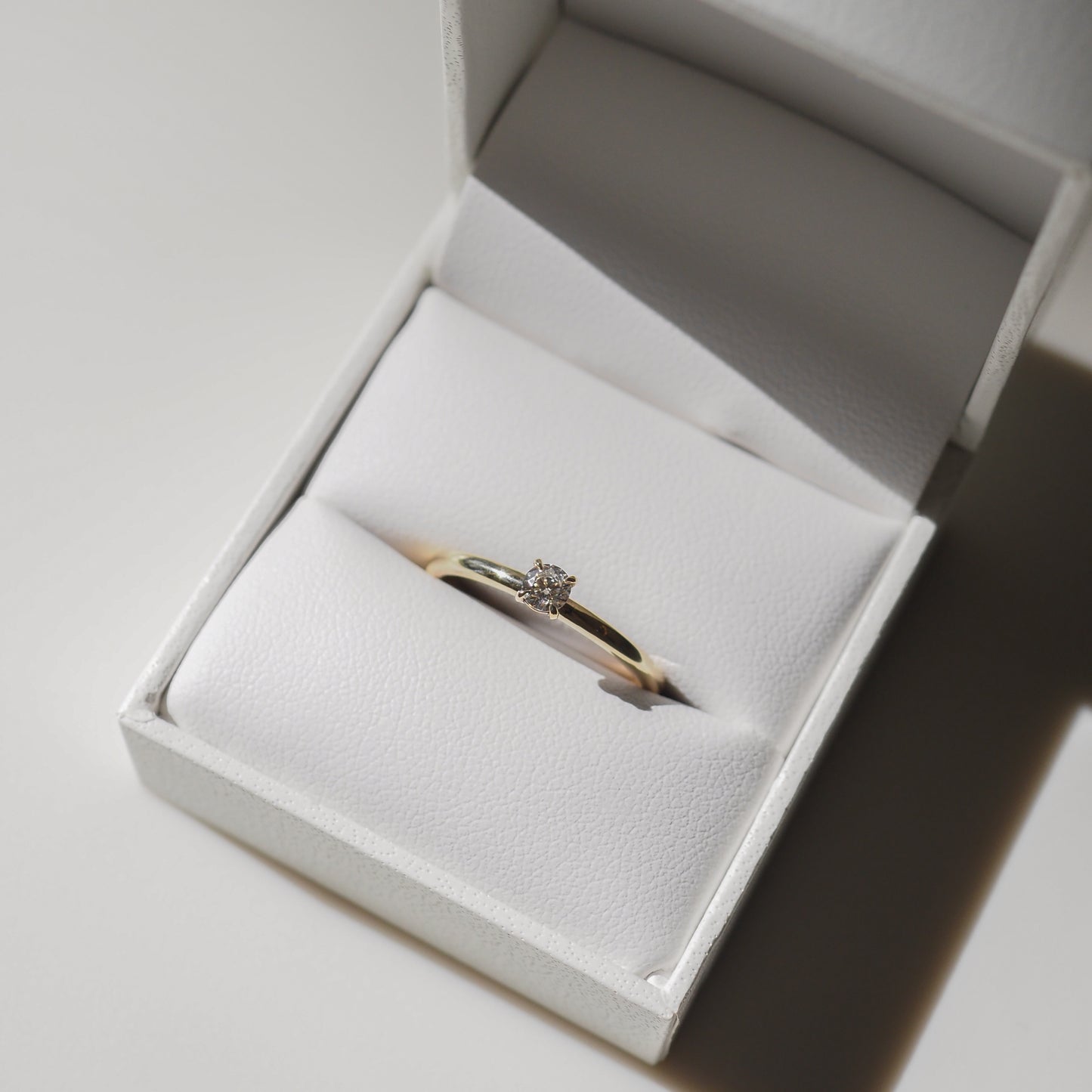 Elegant gold engagement ring with a solitaire diamond in a white leather box, bathed in soft, natural light.