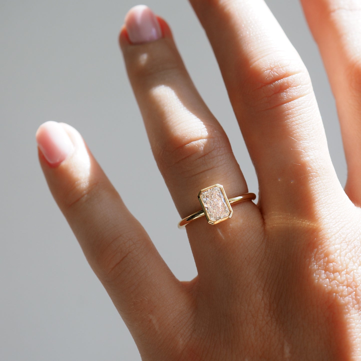 Close-up of a hand wearing a rectangular diamond ring on a gold band. The nails are neatly manicured with pale pink polish.