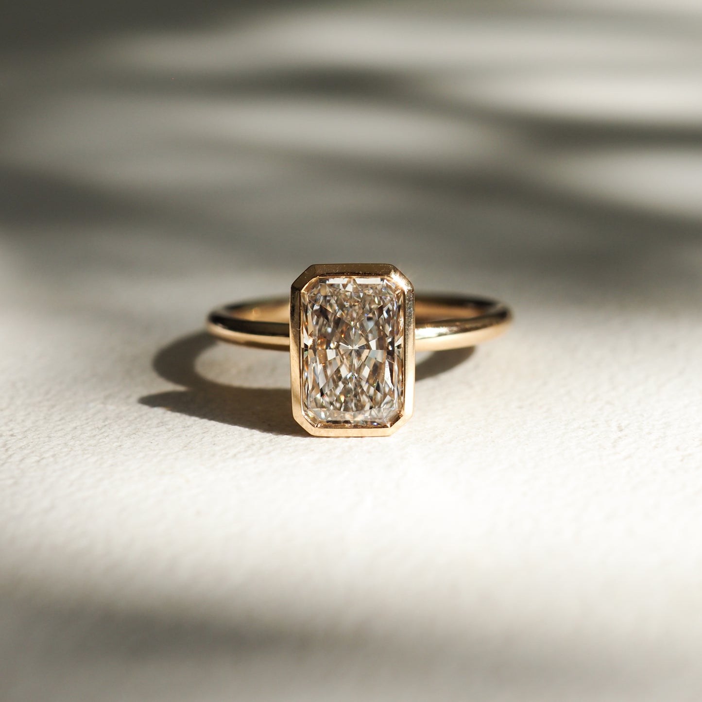 Gold ring with a rectangular, radiant-cut diamond in a minimalist setting, casting a shadow on a textured surface. Luxurious jewellery.