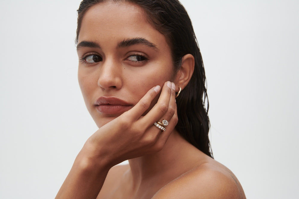 Woman with wet hair wears a diamond ring and stacked bands, looking thoughtfully to the side. Luxury jewellery, minimalist elegance.