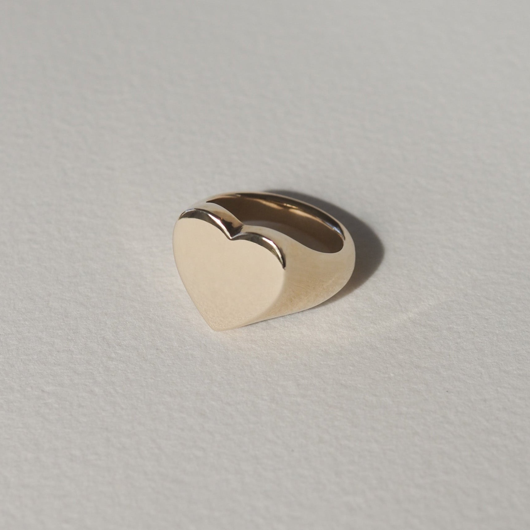 Gold heart-shaped ring on a light textured background, showcasing minimalist luxury jewellery design.