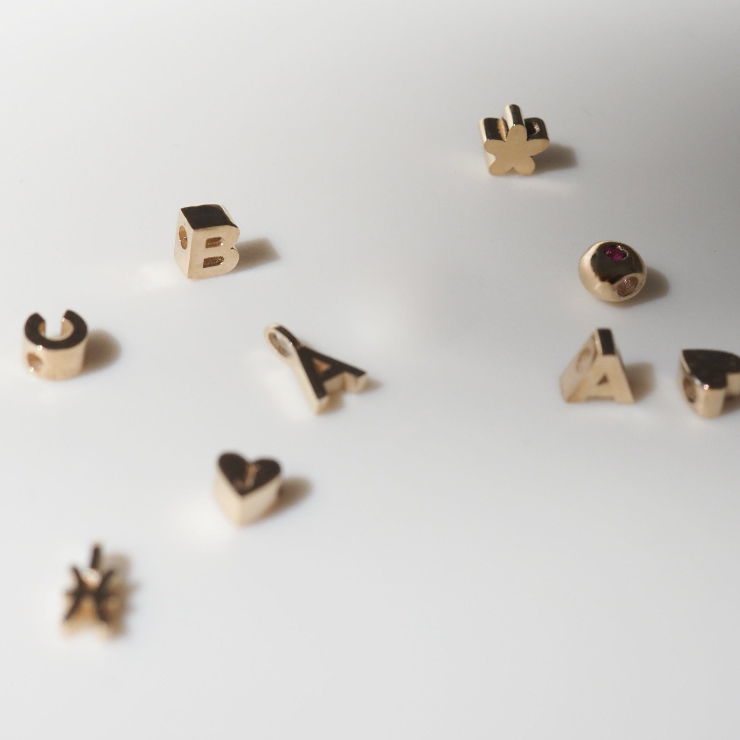 Gold pendant charms in various shapes including letters, a heart, and a star, scattered on a white surface. Luxurious jewellery elements.